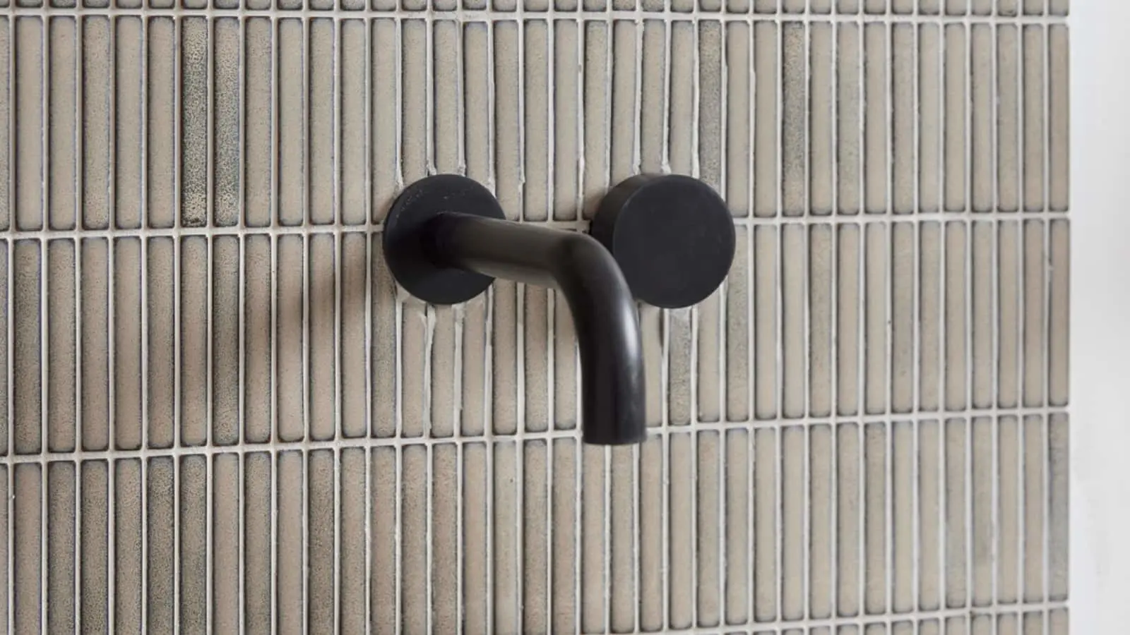 Black wall-mounted faucet with curved spout against beige tiled bathroom wall.