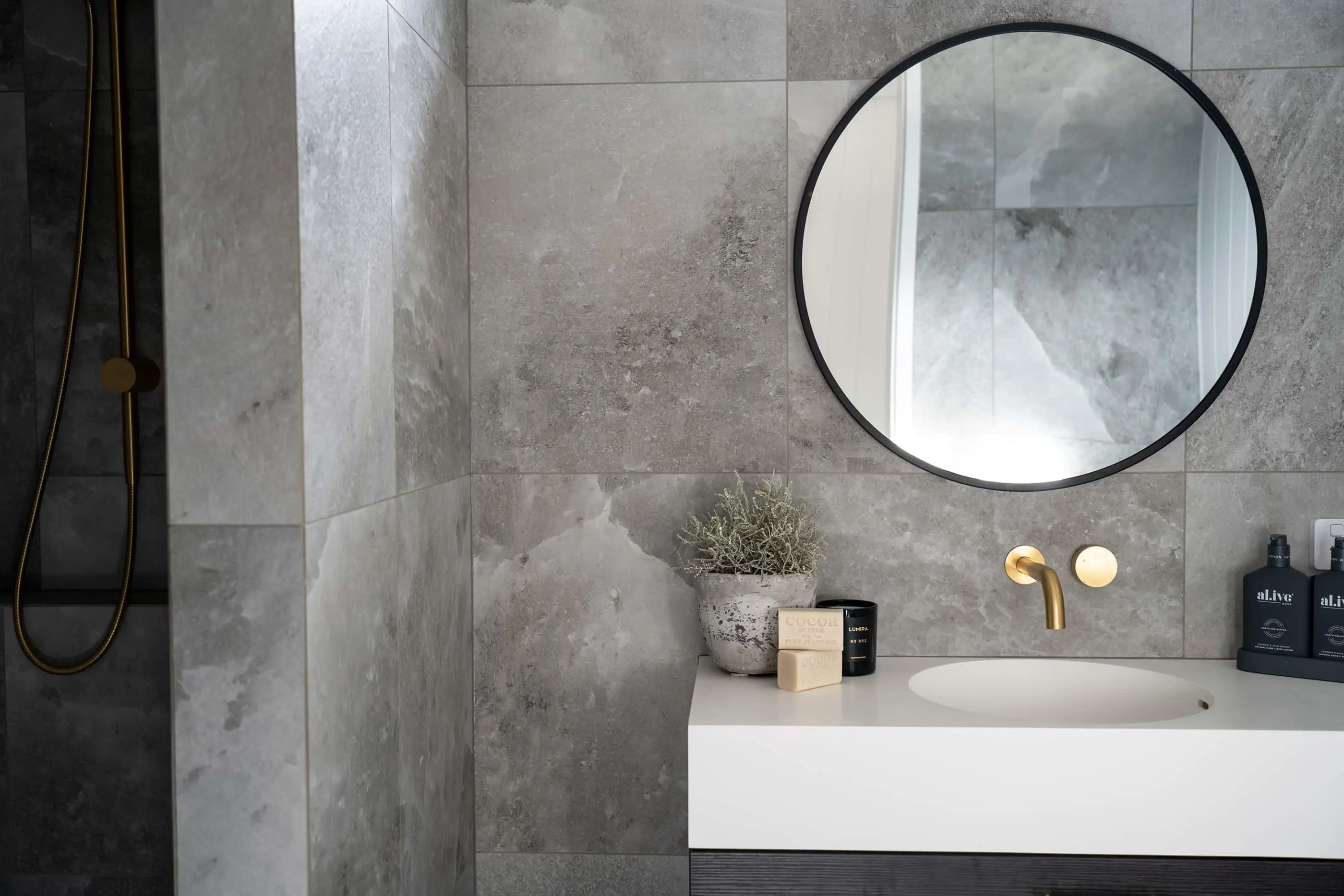 Modern bathroom with gray stone tiles, round black mirror, white sink, gold fixtures, and minimal decorative items.