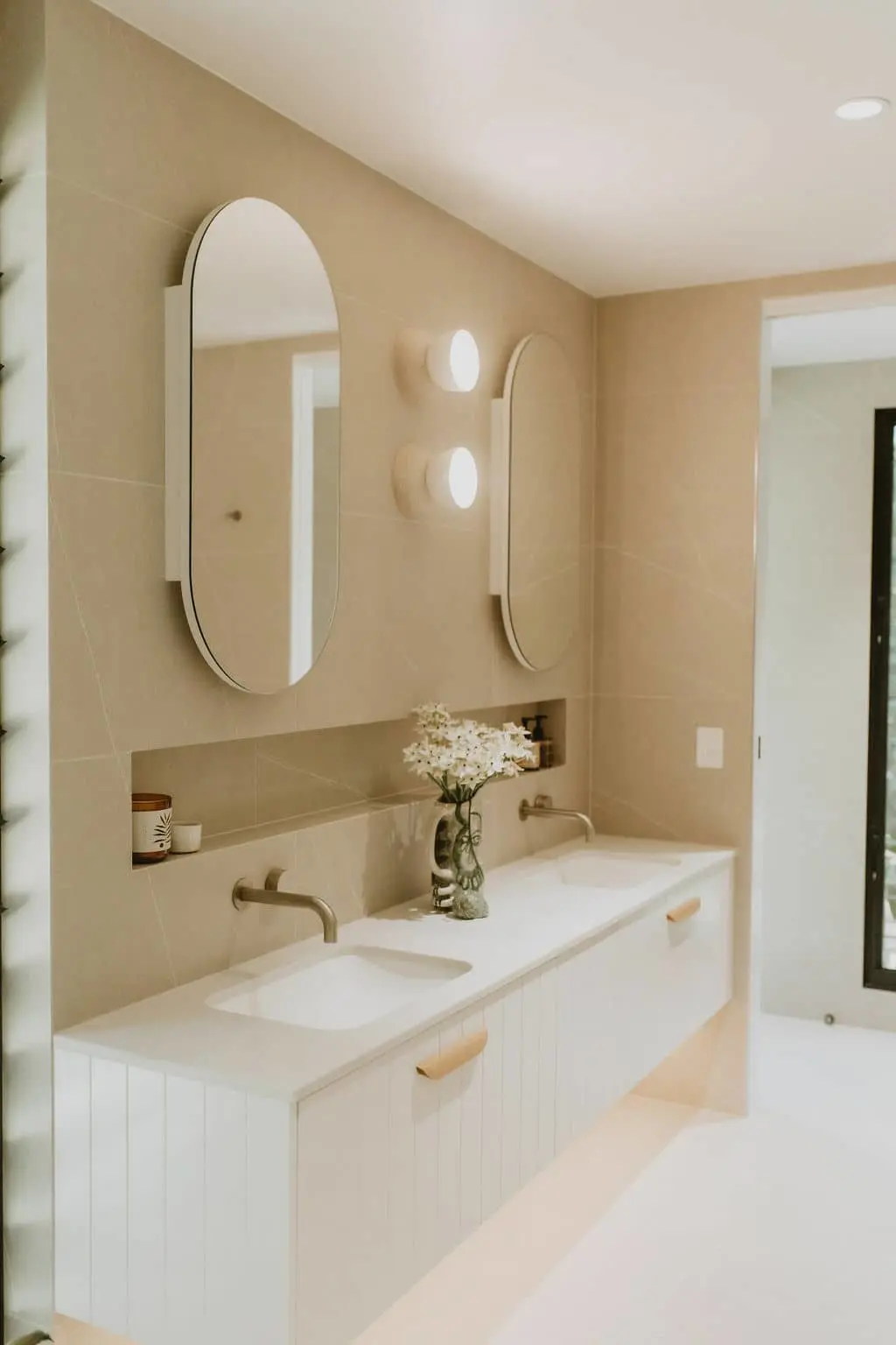 Modern bathroom with beige walls, oval mirrors, white vanity with double sinks, and small vase of white flowers.