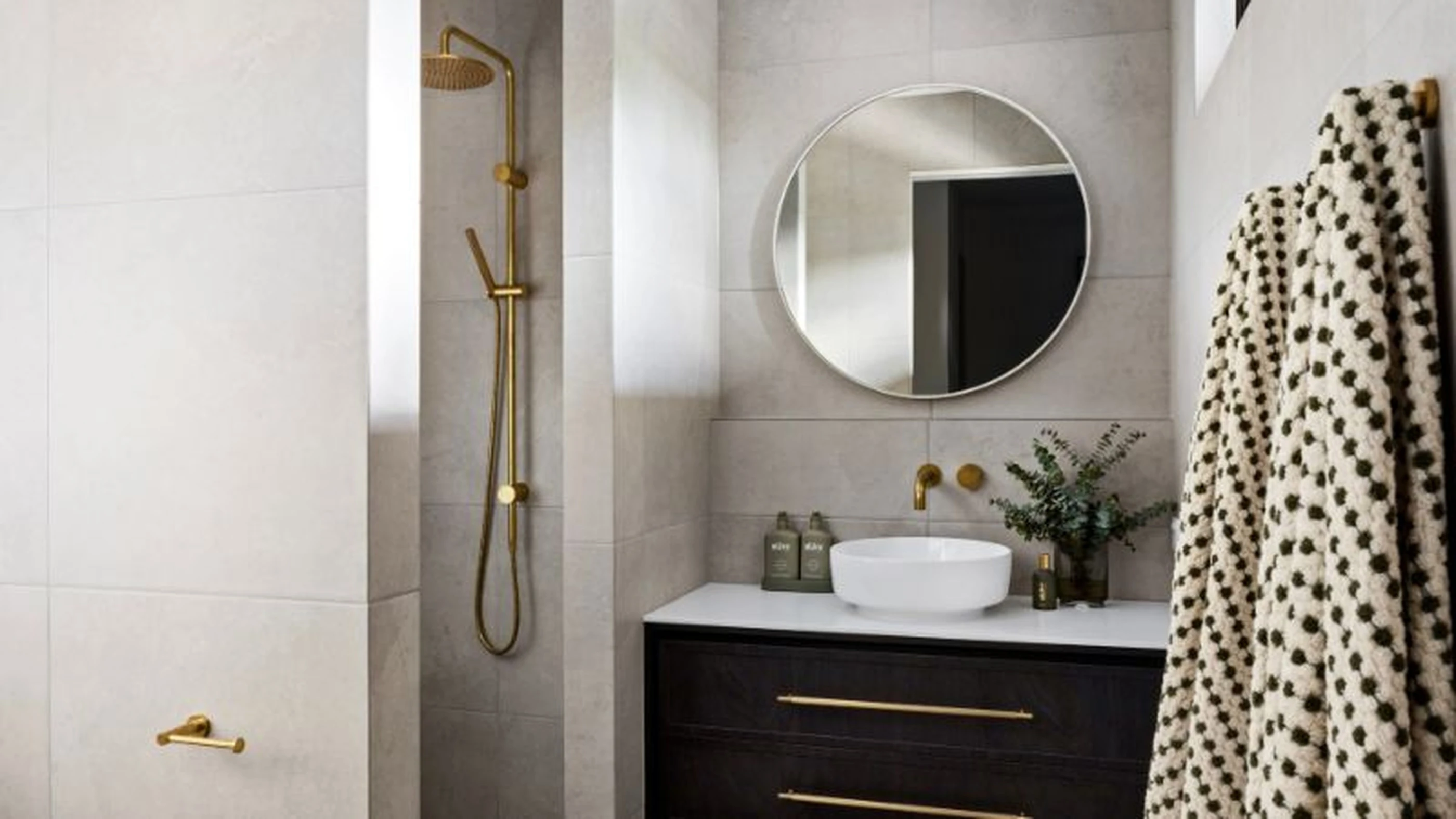 Modern bathroom with gray tiles, gold fixtures, round mirror, white vessel sink, and black-spotted white towel.