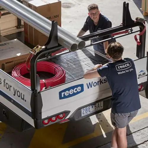 Two workers loading metal pipes and red coiled material onto a Reece utility truck in a warehouse.