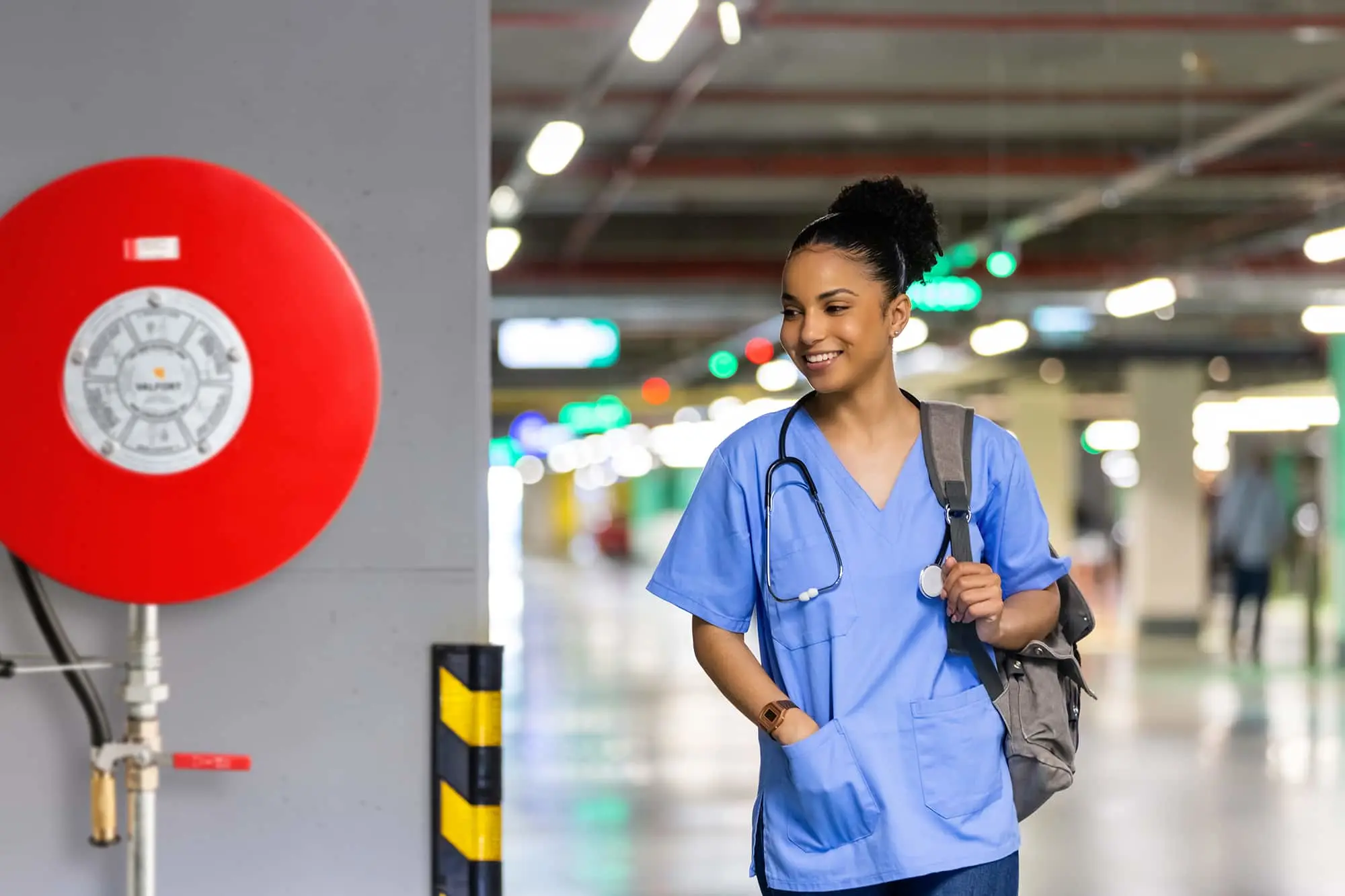 Healthcare worker in blue scrubs with stethoscope walking through parking garage, red fire alarm visible on wall.