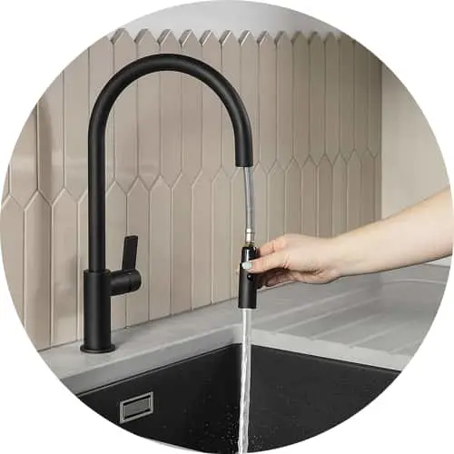 Hand pulling out extendable nozzle of black kitchen faucet with water running into sink against beige tile backsplash.