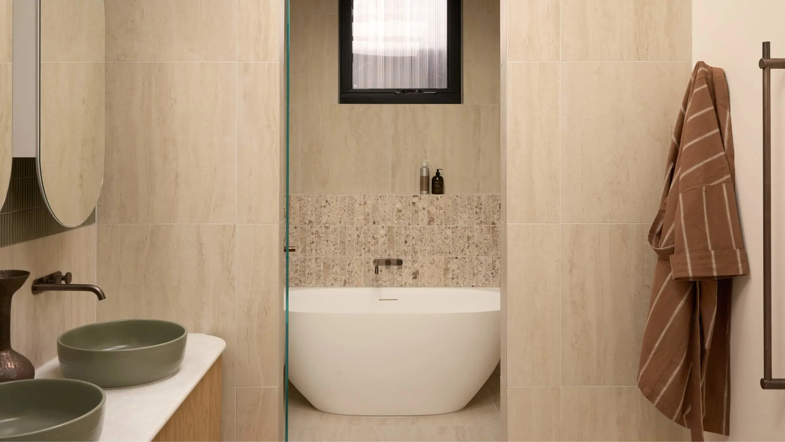 Modern bathroom with beige tiles, white freestanding tub, green vessel sinks, glass shower, and brown striped towel.