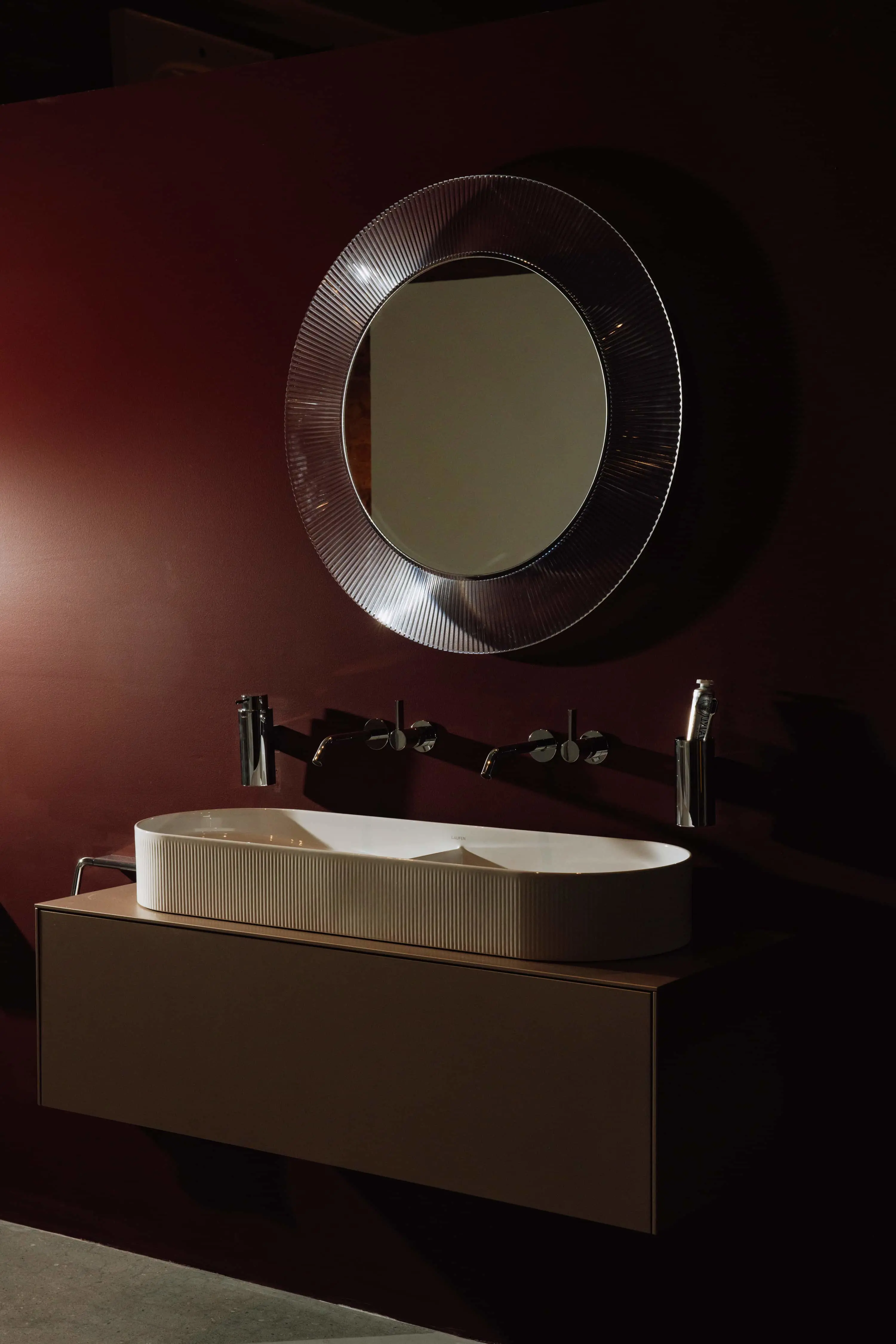 Modern bathroom with white vessel sink on wooden vanity, circular mirror with textured frame, and burgundy walls.