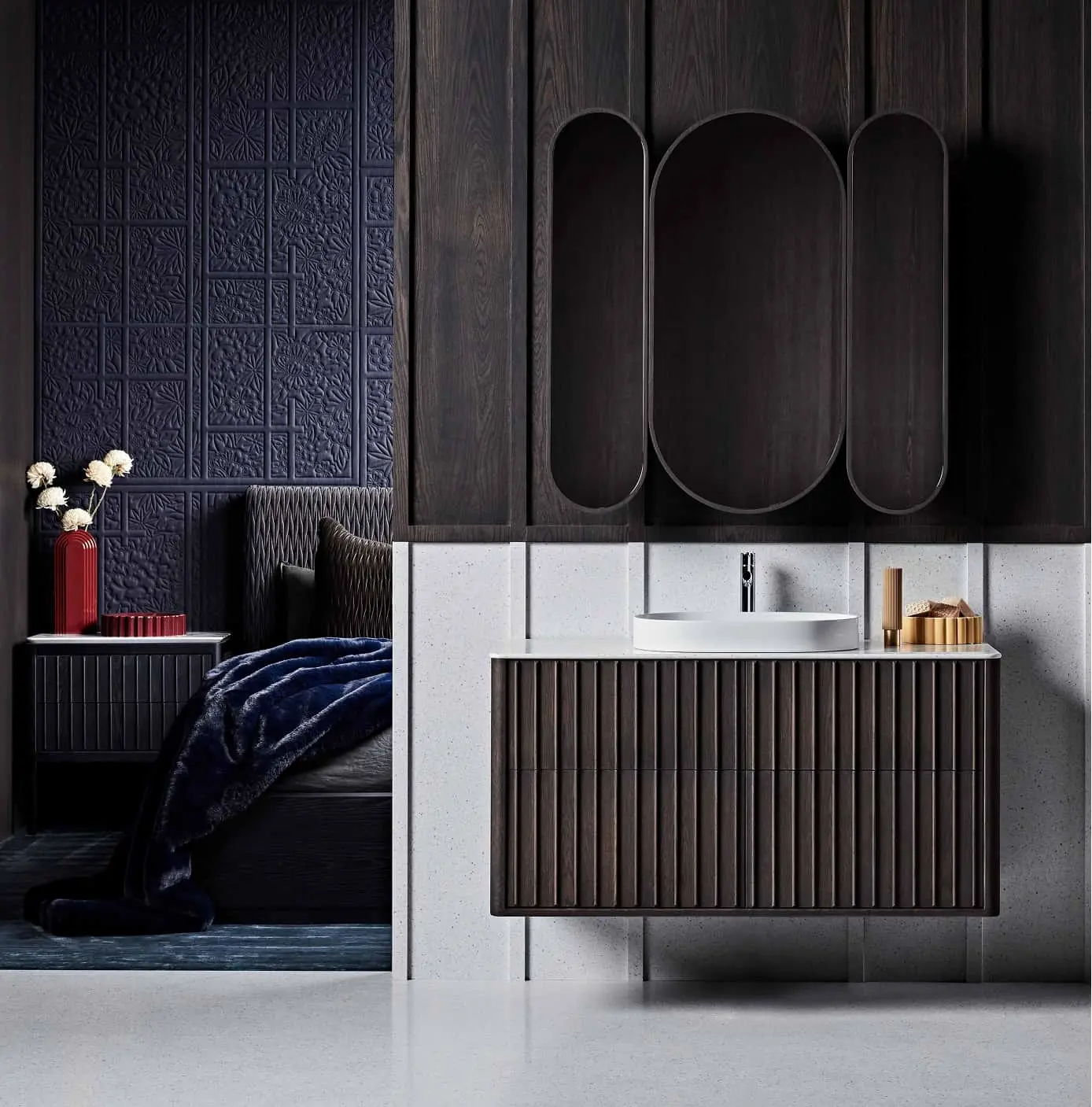 Modern bathroom with dark wood vanity, white sink, textured navy wall, and glimpse of bedroom with red vase and blue throw.