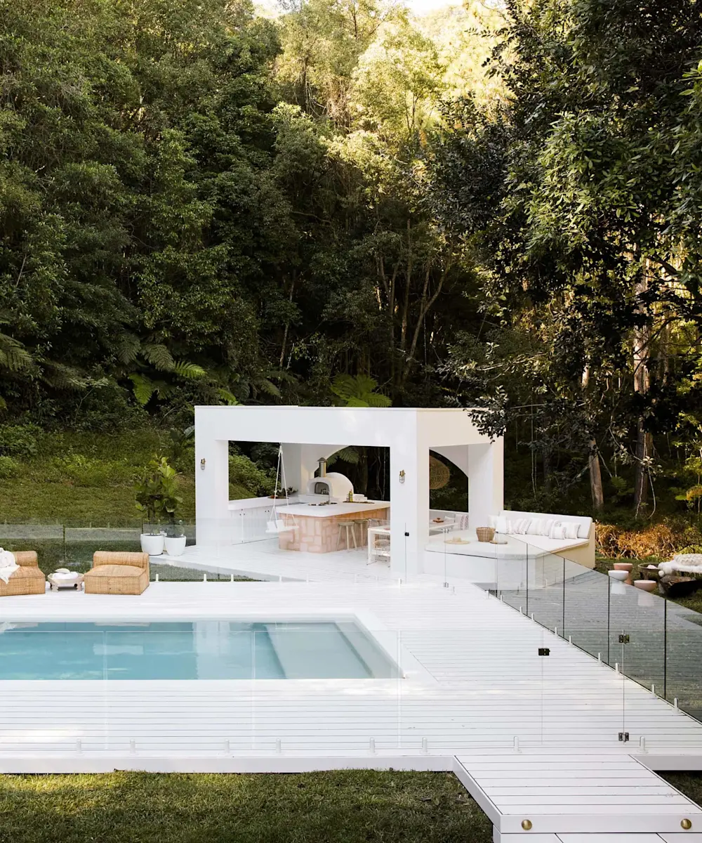Modern white outdoor kitchen and lounge area with swimming pool, surrounded by lush forest greenery.
