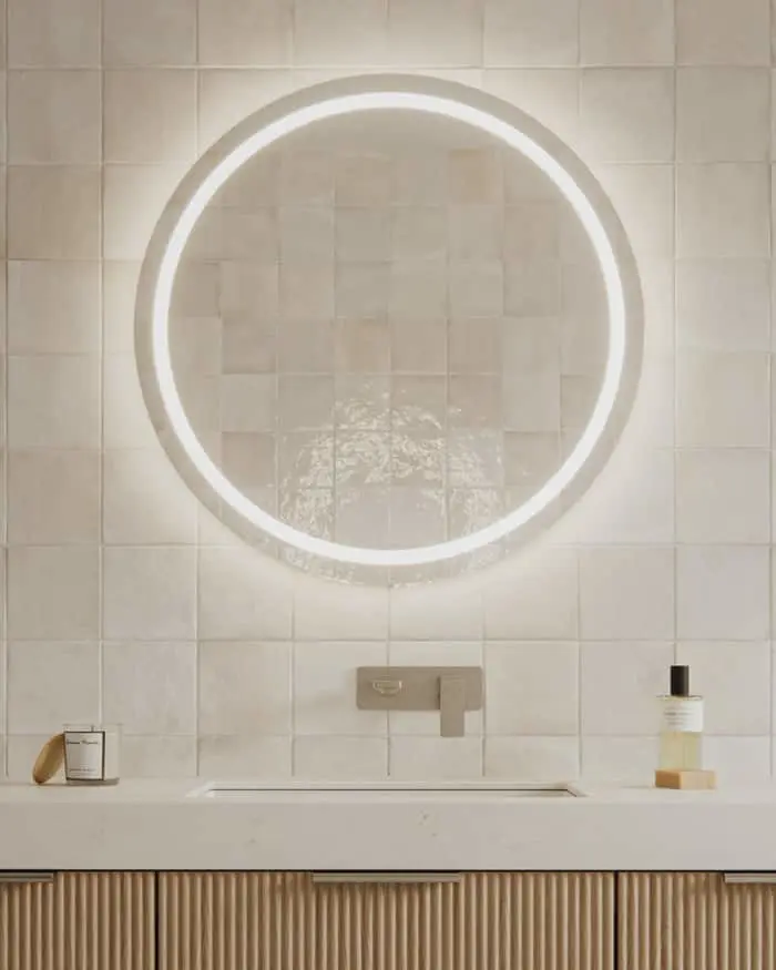 Modern bathroom with round backlit mirror above sink, beige tiled wall, and wooden cabinet below.