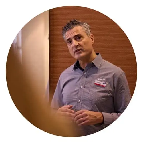 A person in a gray button-up shirt with a name tag speaking in front of a brick wall background.