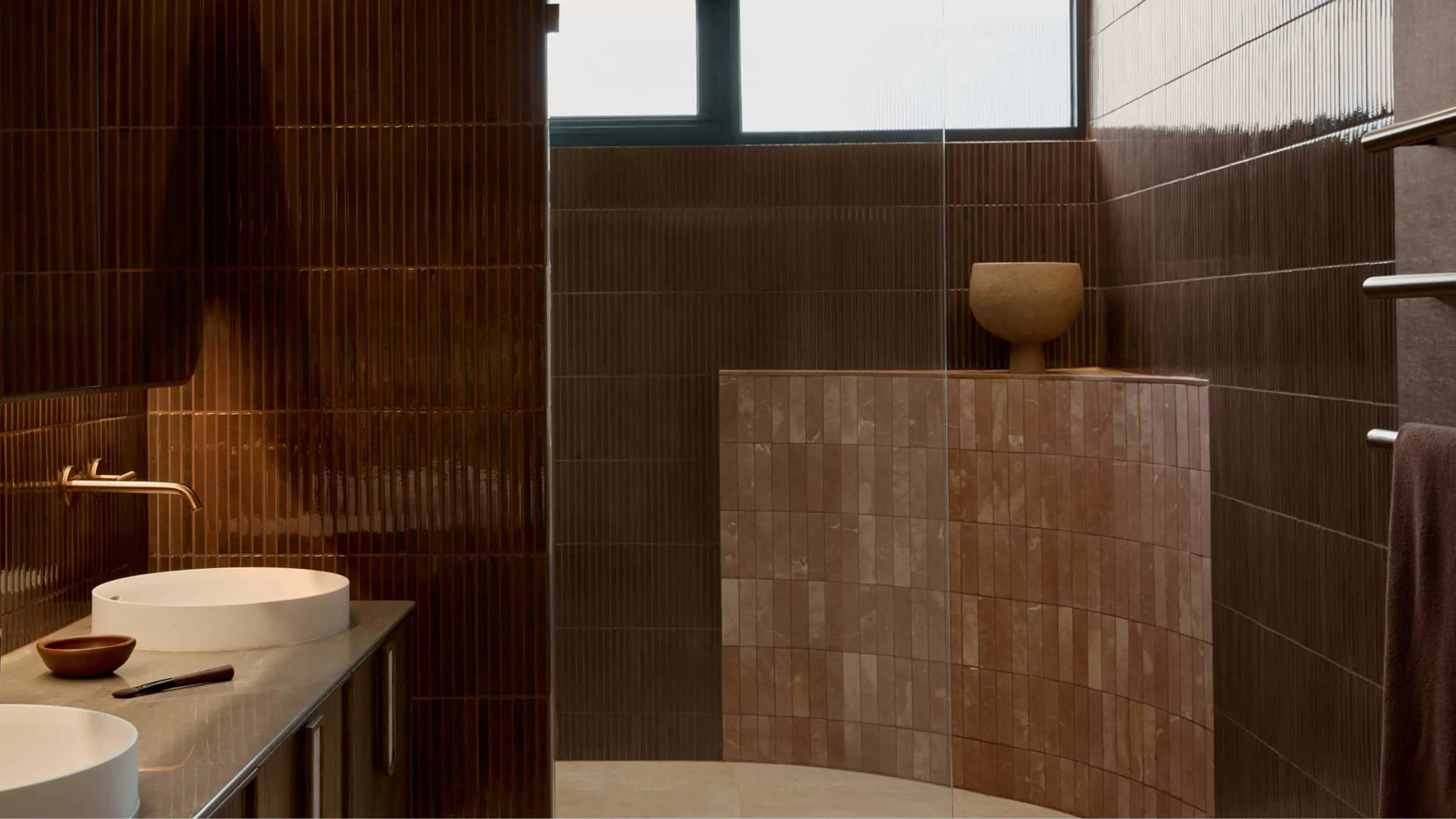 Modern bathroom with brown tiled walls, white basin sinks, and a curved partition with decorative ceramic vessel.
