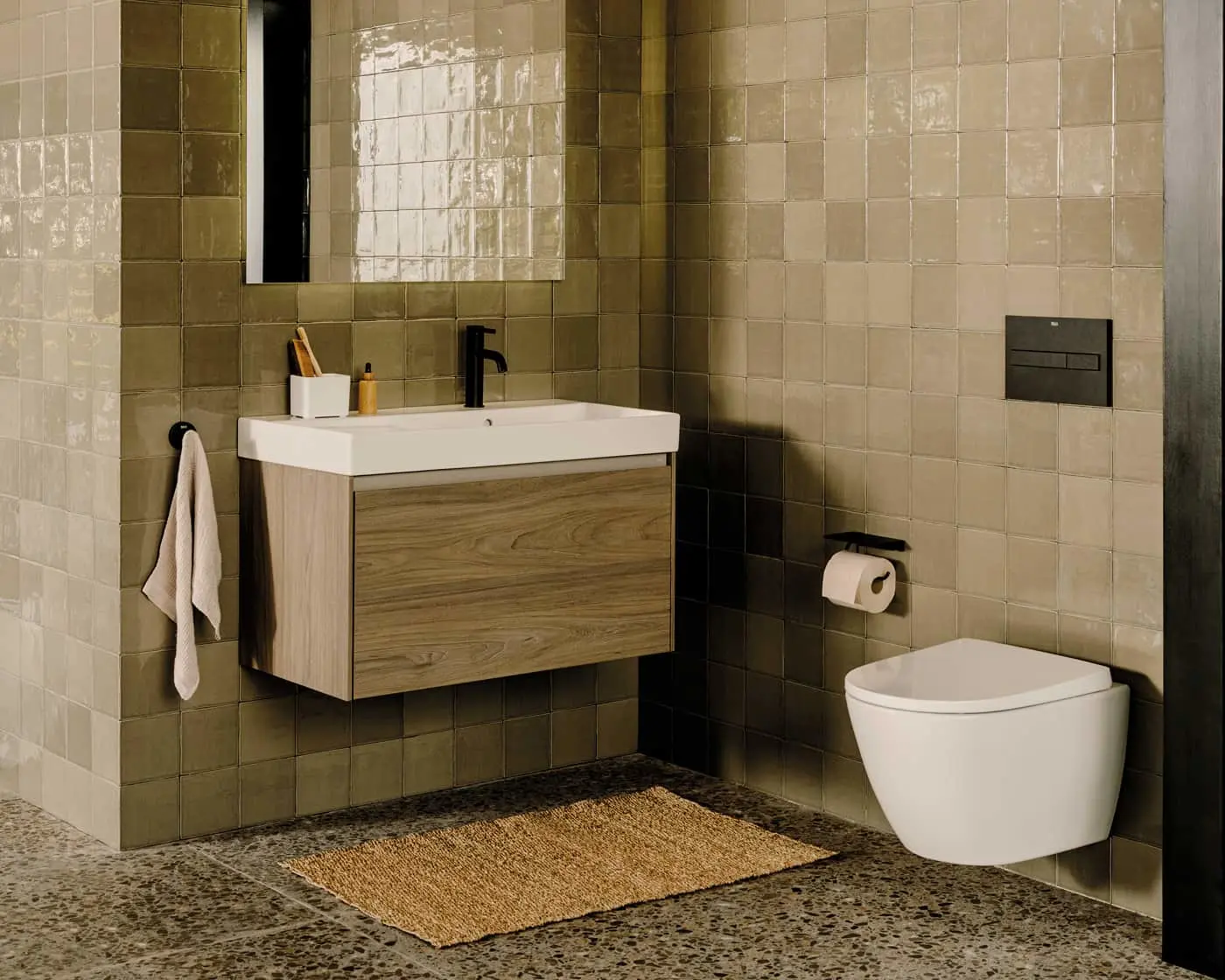 Modern bathroom with olive green tile walls, wooden vanity with white sink, wall-mounted toilet, and jute rug on stone floor.
