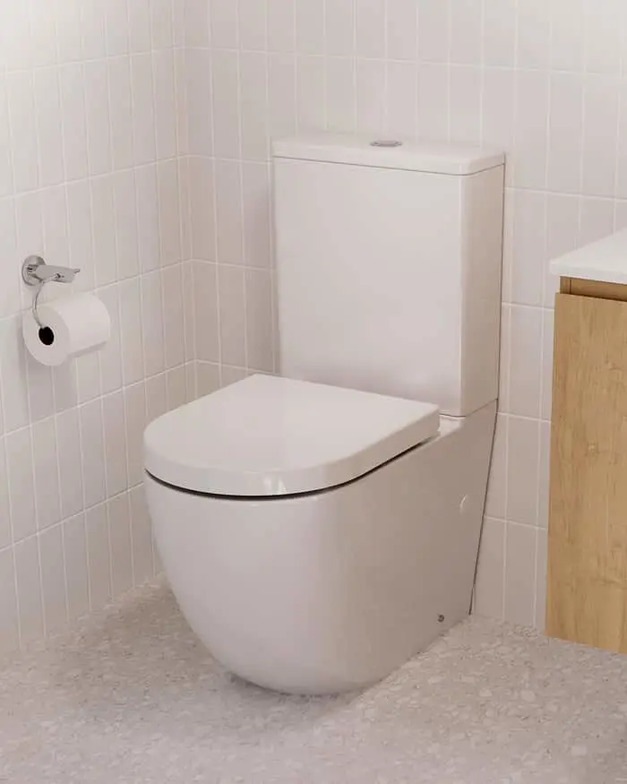White toilet with closed lid in a bathroom with white tiled walls, toilet paper holder, and wooden cabinet edge visible.