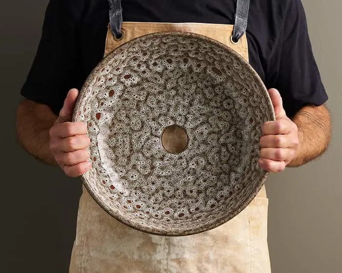 Person in apron holding a large ceramic bowl with speckled glaze pattern and center hole.