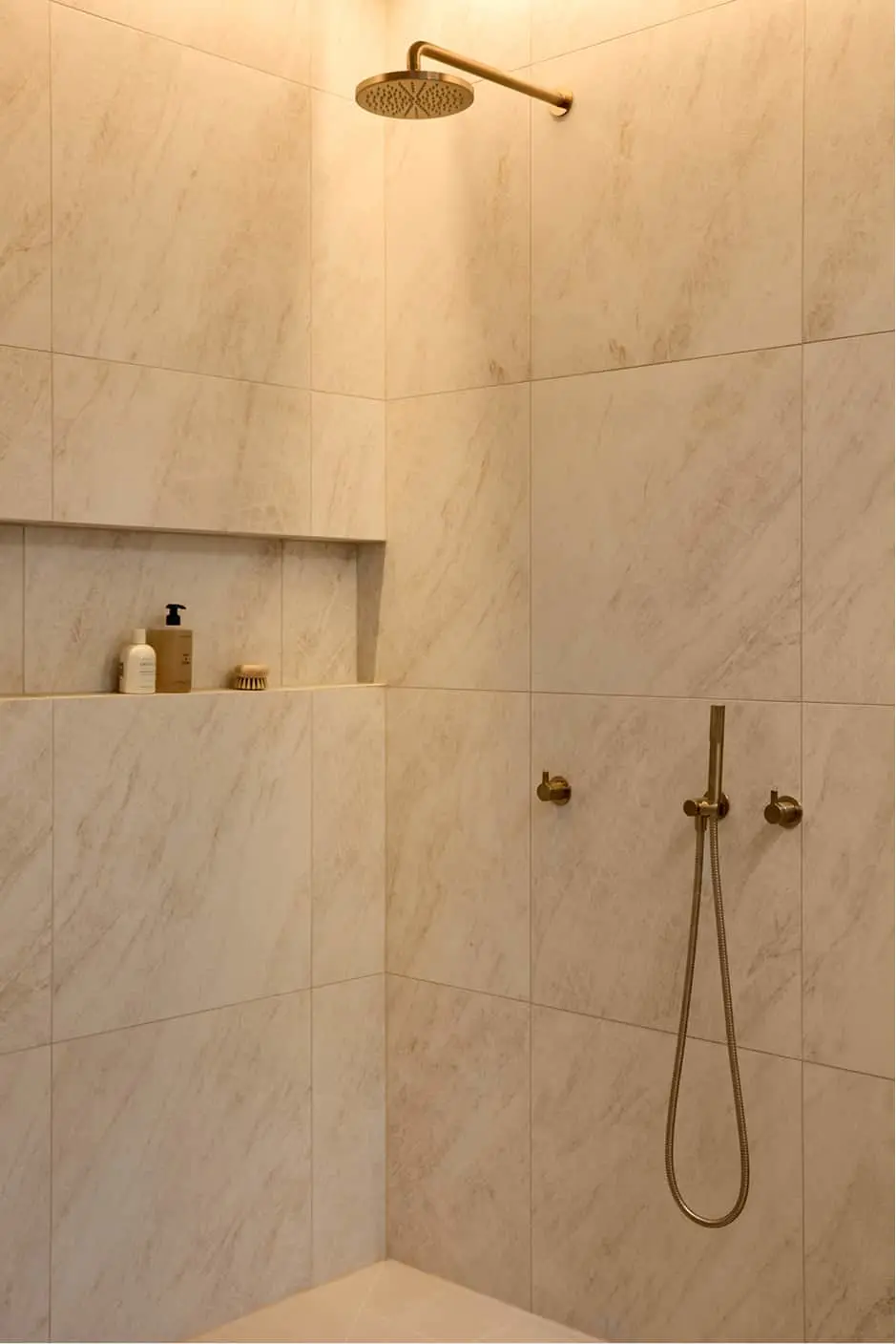 Walk-in shower with cream marble tiles, brass overhead rain shower, handheld shower, and a recessed niche holding toiletries.