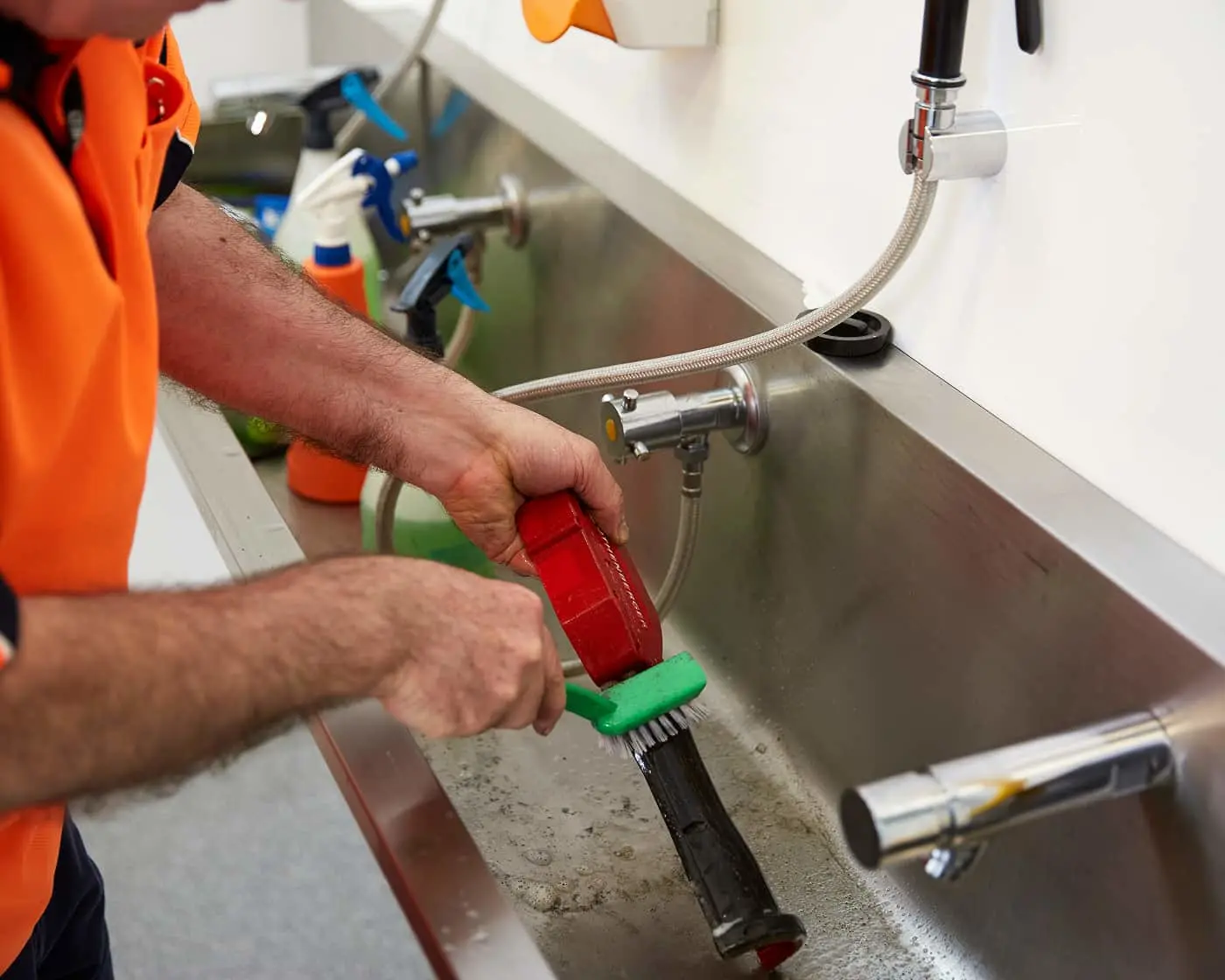 Plumber in orange shirt using a red and green brush tool to clean a kitchen sink drain, with cleaning supplies nearby.