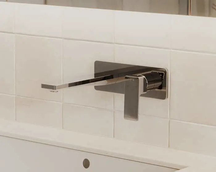 Modern wall-mounted bathroom faucet with brushed metal finish against white tile backsplash.