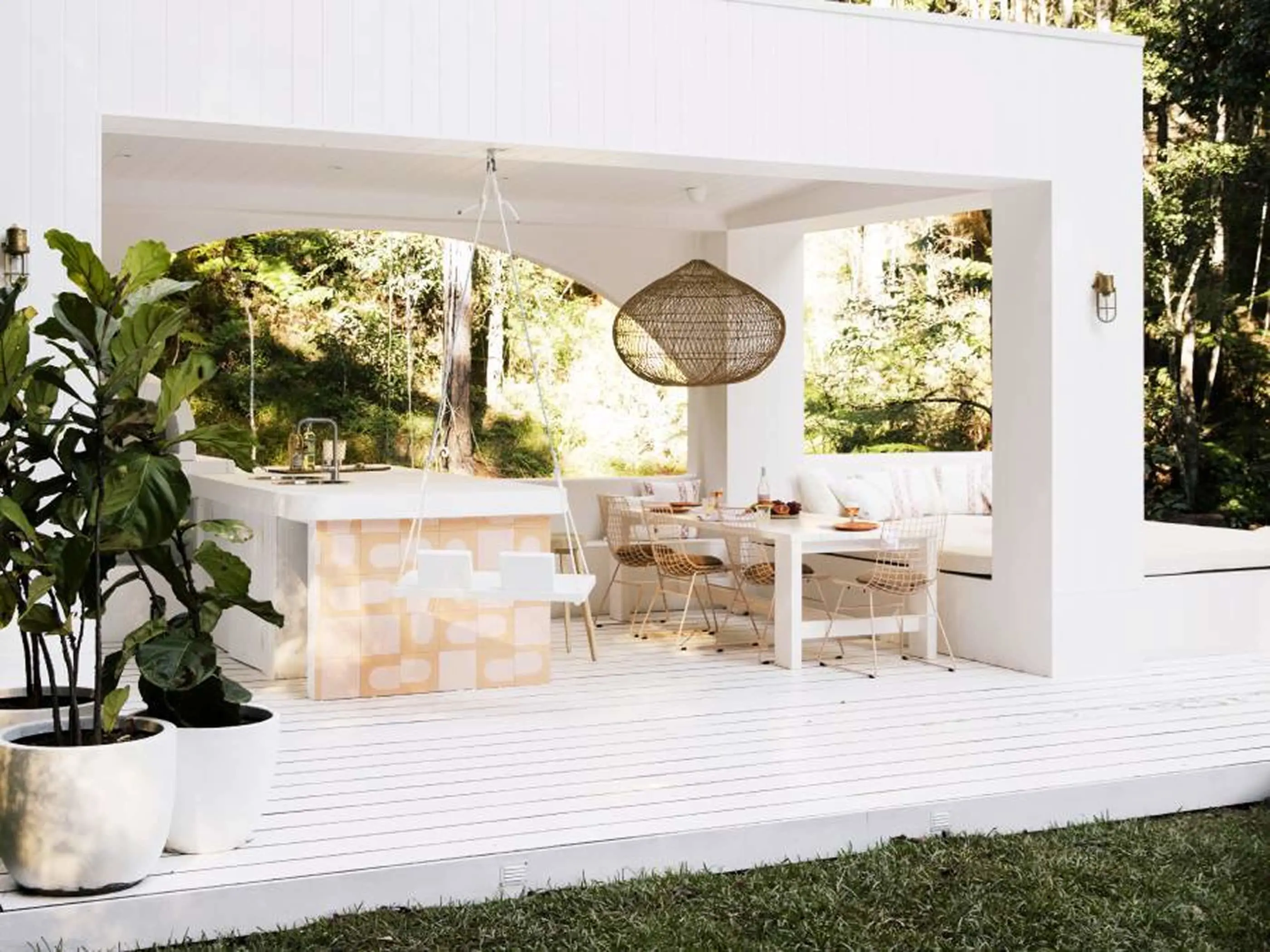 Modern white outdoor patio with dining area, wicker pendant light, and bar counter surrounded by lush greenery.