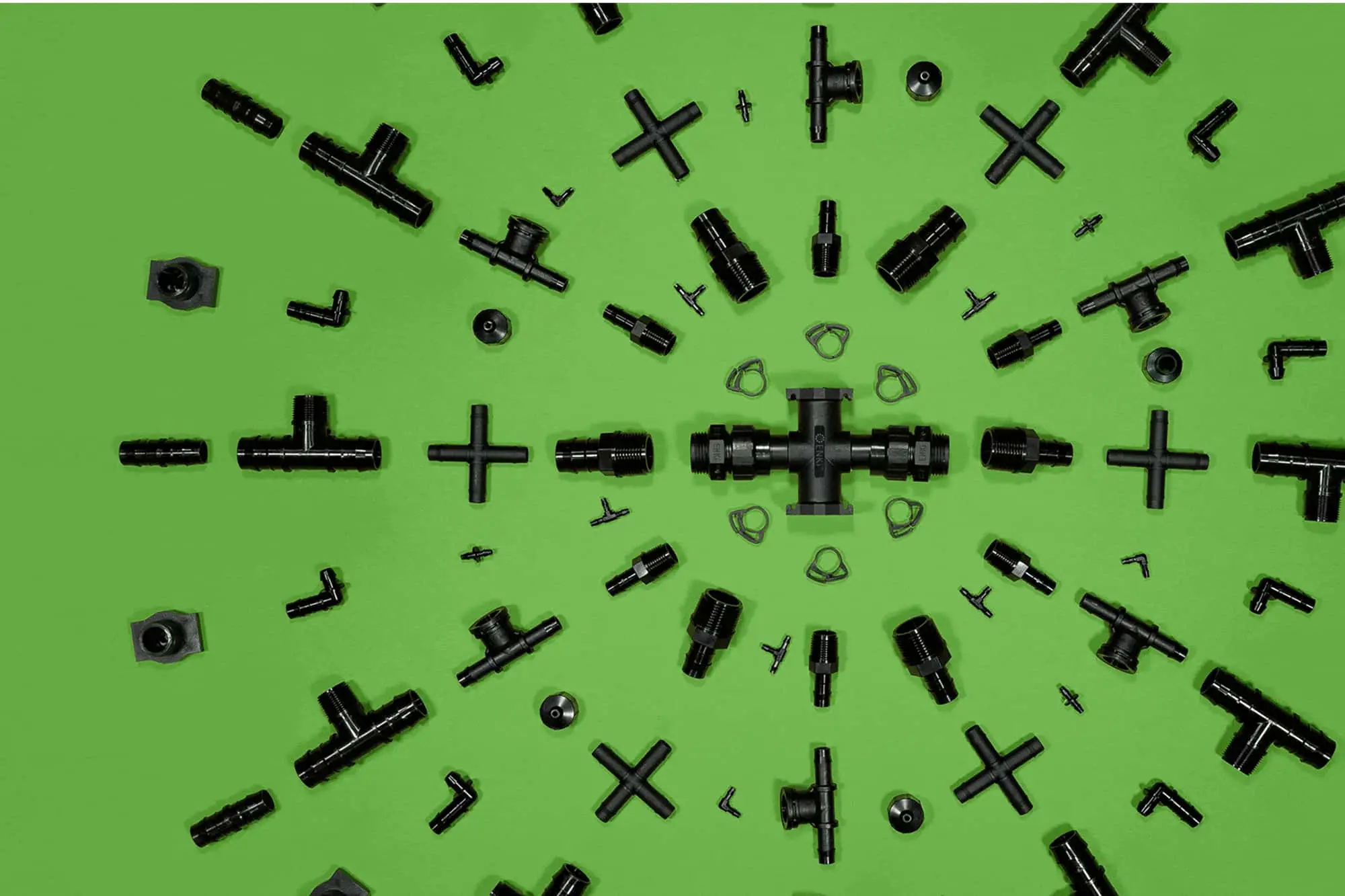 Black plumbing or irrigation fittings arranged on bright green background, featuring T-joints, connectors, and cross pieces.