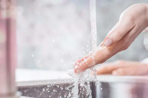 Hand washing under running water with splashing droplets in a bathroom sink.