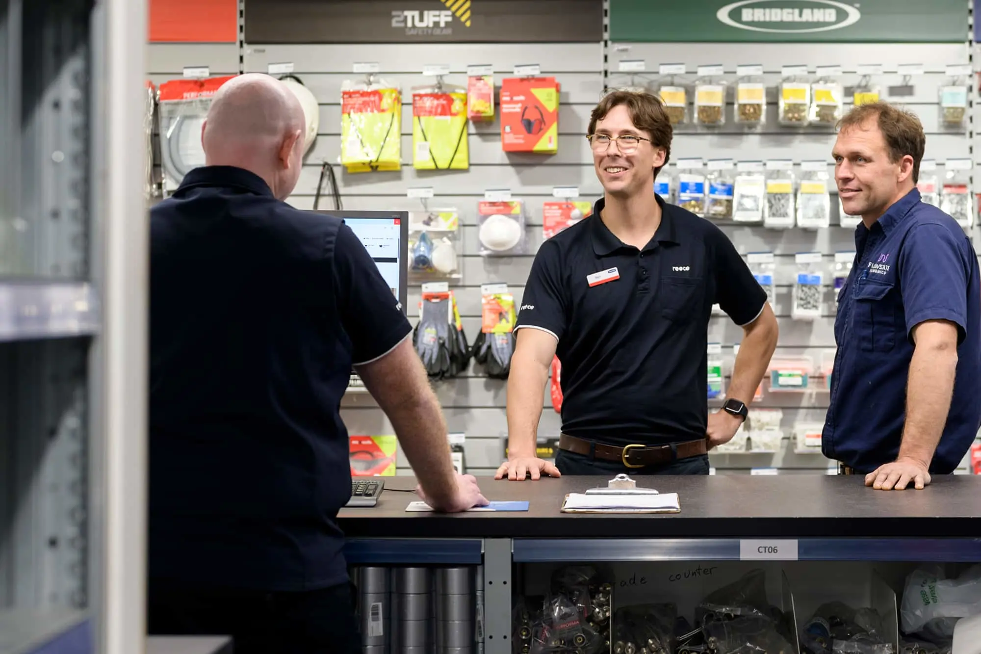 Three men at a hardware store counter with supplies displayed on wall shelves behind them.