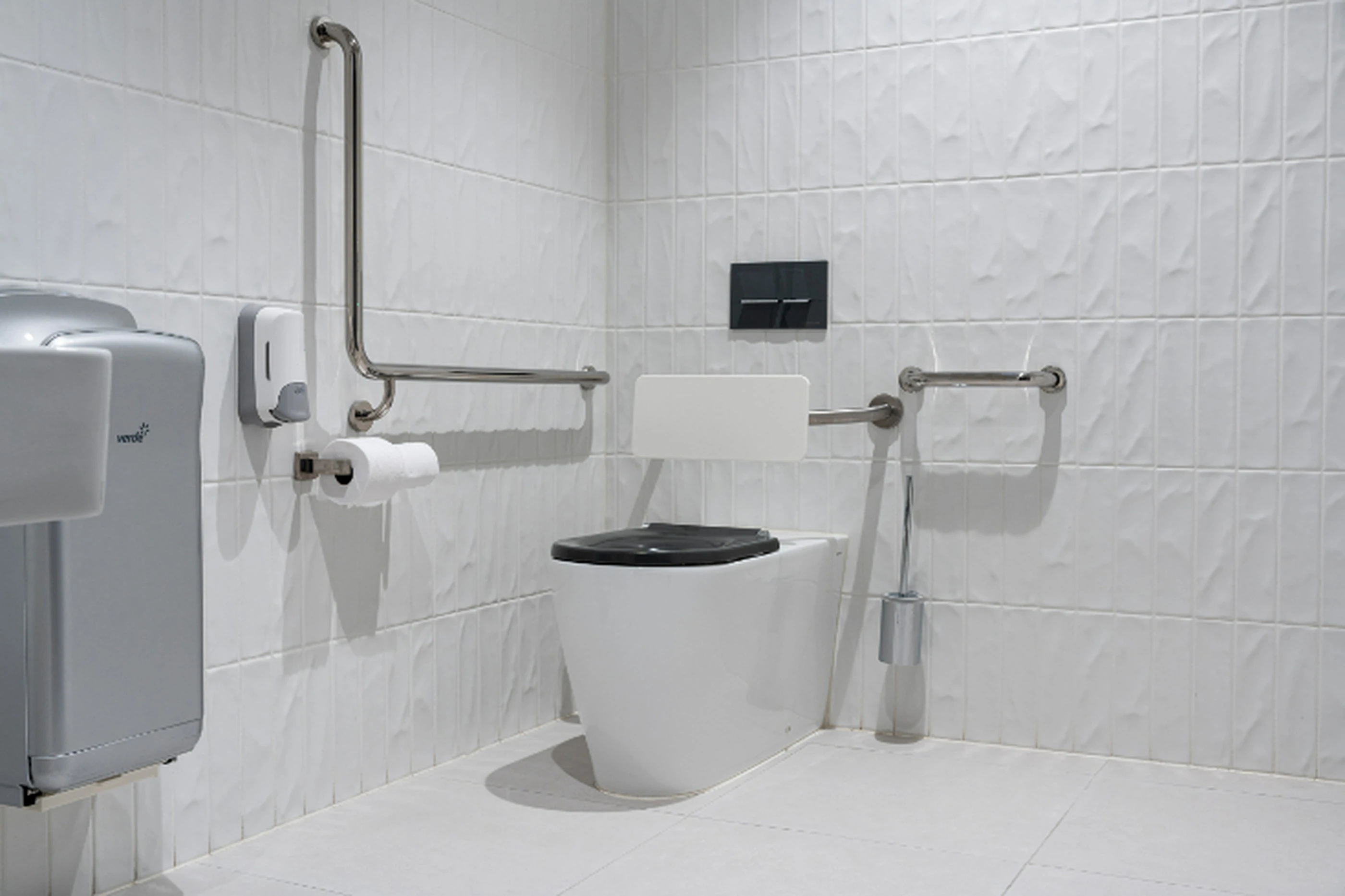 Accessible bathroom with white toilet, metal grab bars, toilet paper dispenser, and toilet brush in white tiled space.