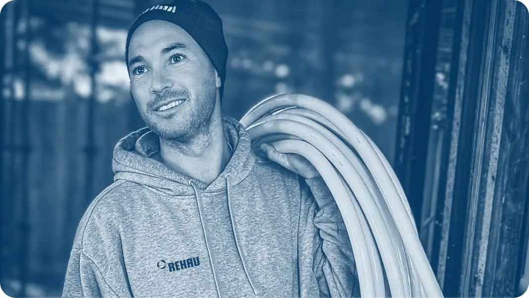 Person in beanie and REHAU hoodie carrying coiled white pipes over shoulder in blue-tinted construction setting.