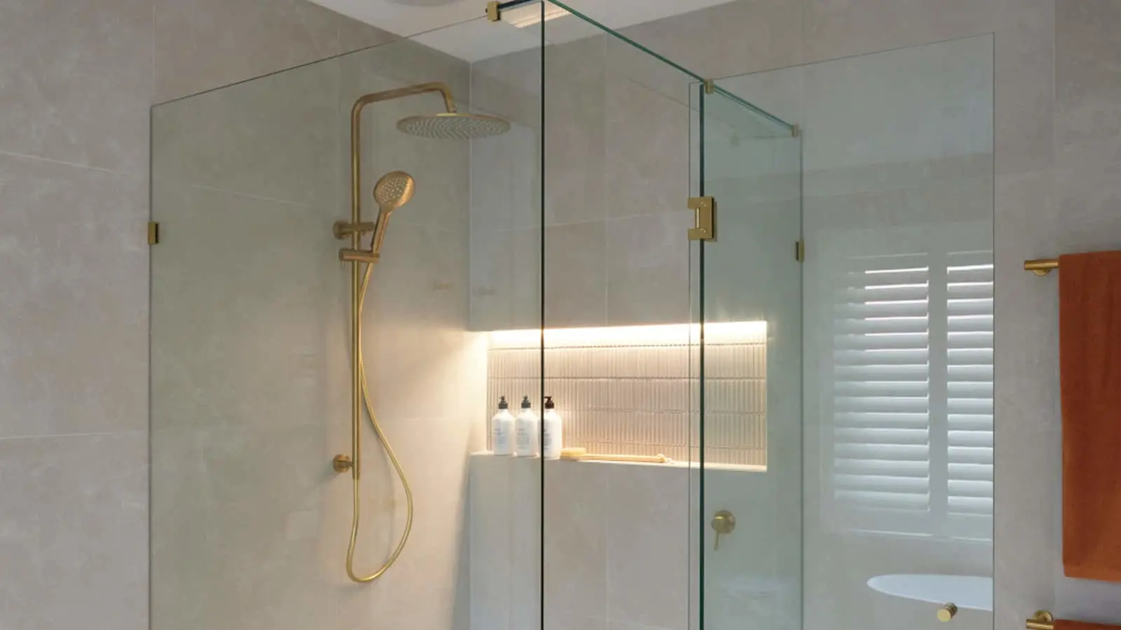 Modern glass shower enclosure with gold fixtures, illuminated niche for toiletries, and white tiled walls.