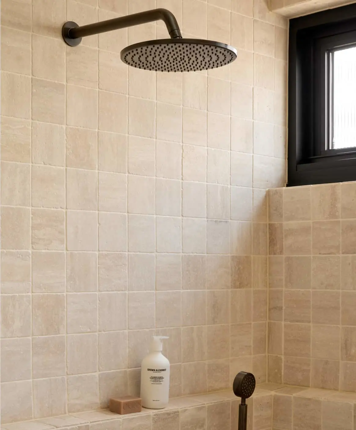 Modern shower with beige tile walls, gunmetal rainfall showerhead, small window, and built-in shelf with soap bottle.
