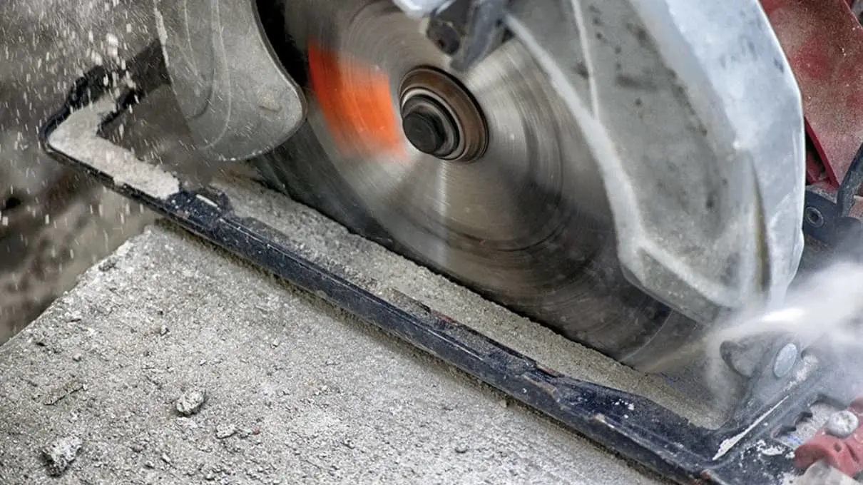 Circular saw blade in motion cutting through concrete, creating dust and debris with a glowing orange edge from friction.