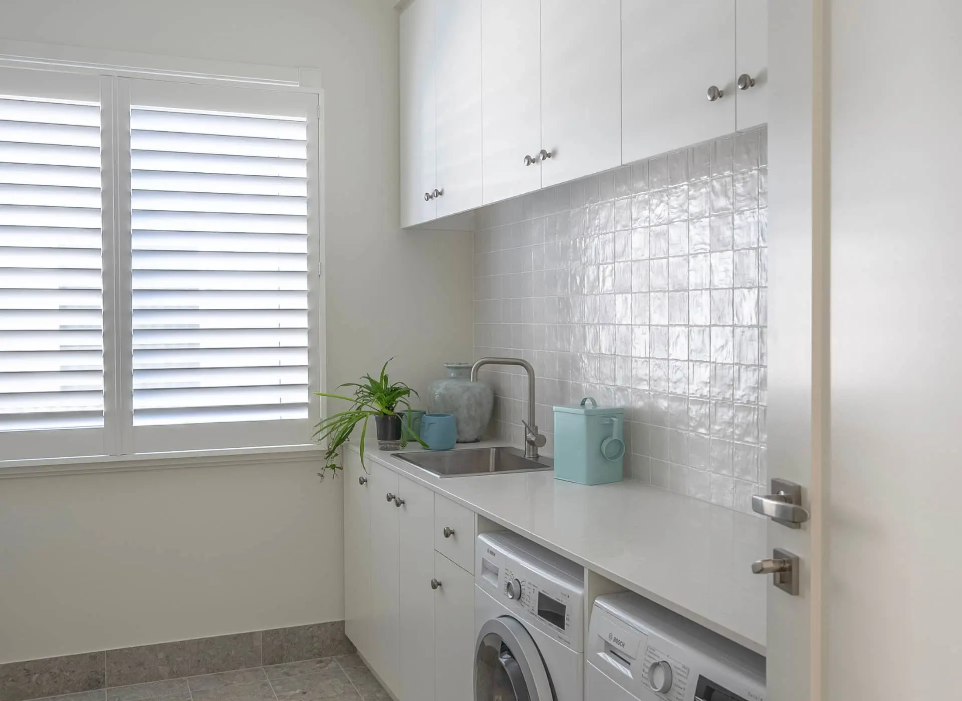 Modern white laundry room with sink, washing machines, shiny tile backsplash, window shutters, and small plant.