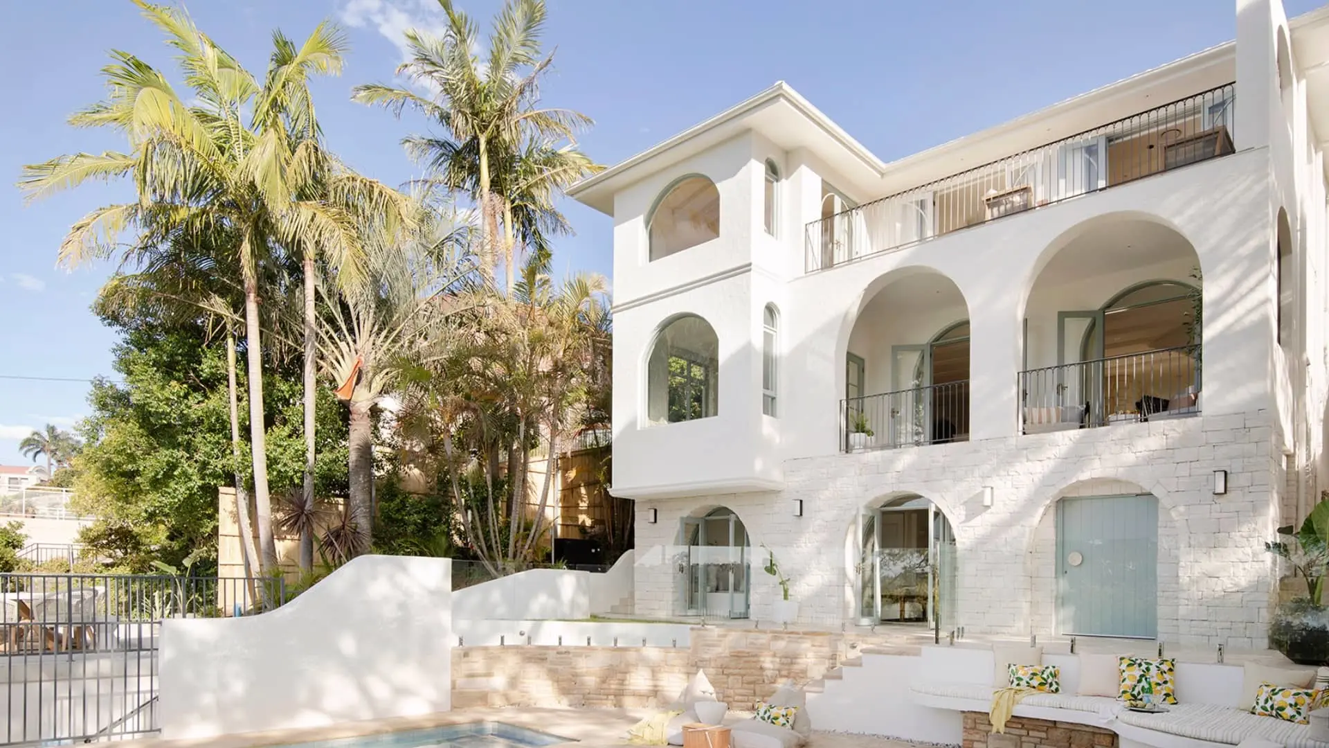 Modern white Mediterranean-style villa with arched windows, balconies, palm trees, and poolside seating area under blue sky.