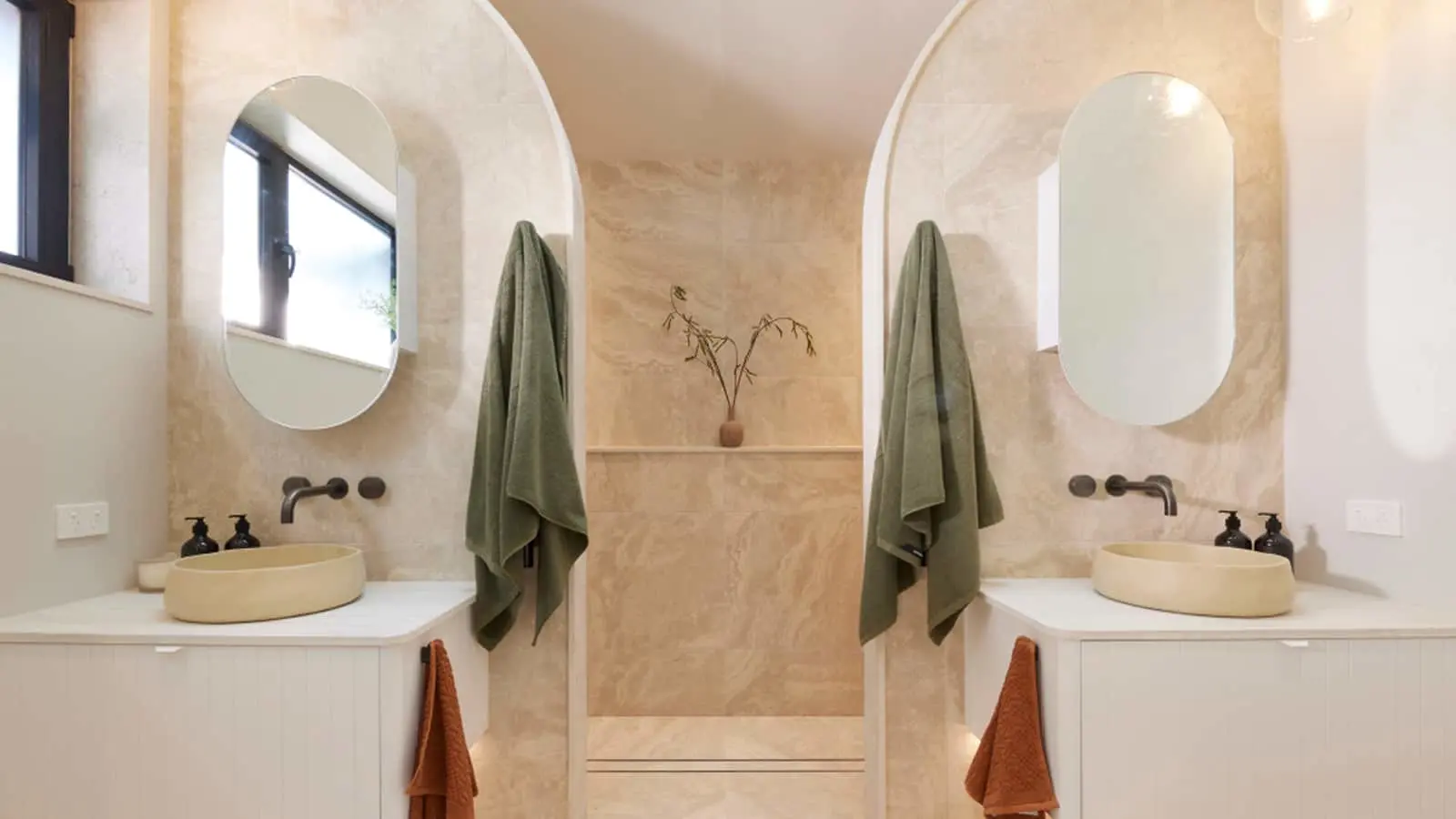 Modern bathroom with dual cream sinks, oval mirrors, green towels, and beige textured walls featuring a small plant.