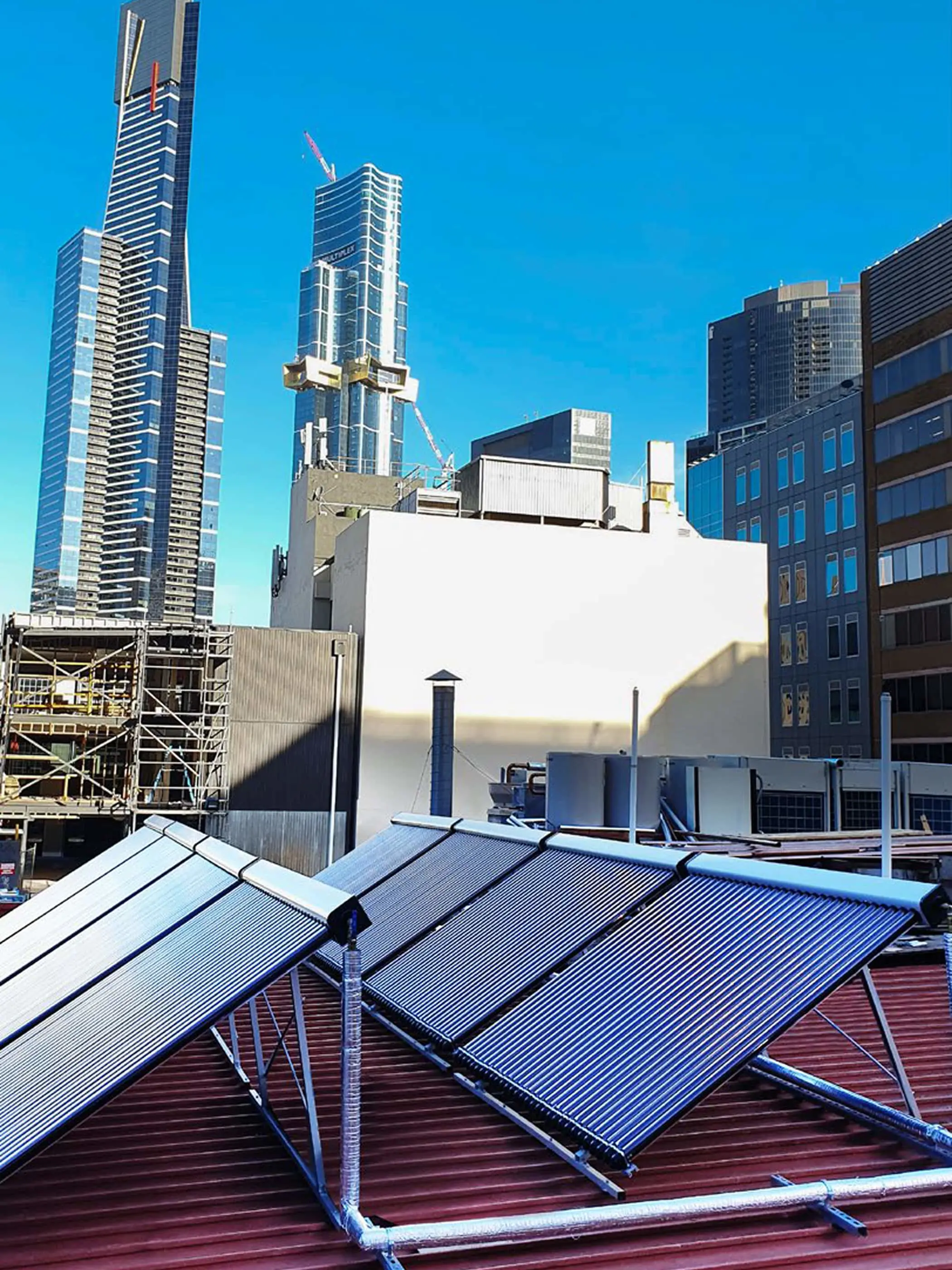 Solar panels installed on a rooftop with modern glass skyscrapers and construction cranes against a clear blue sky.