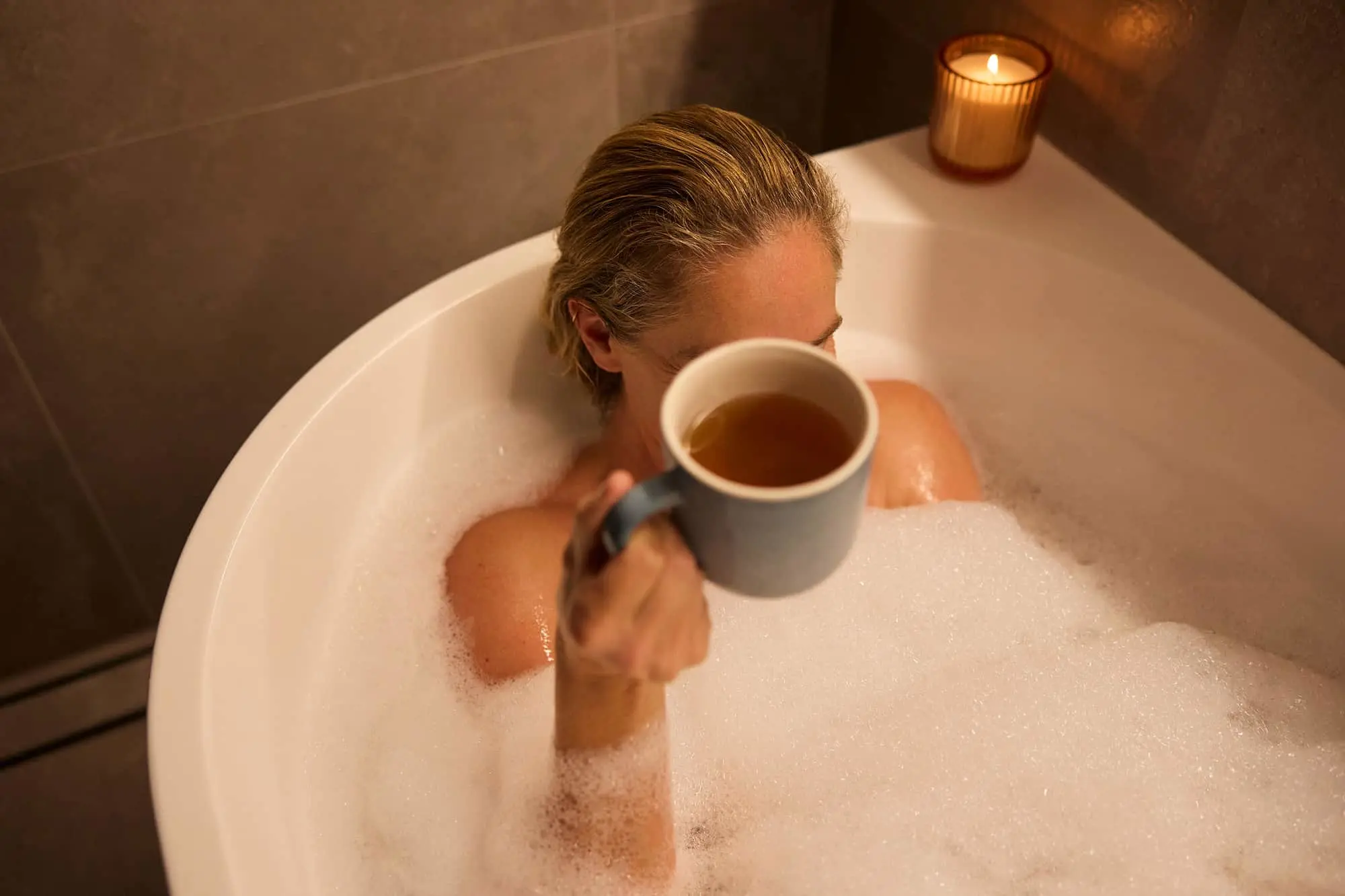 Person relaxing in a bubble bath holding a mug of tea, with a lit candle on the edge of the tub.