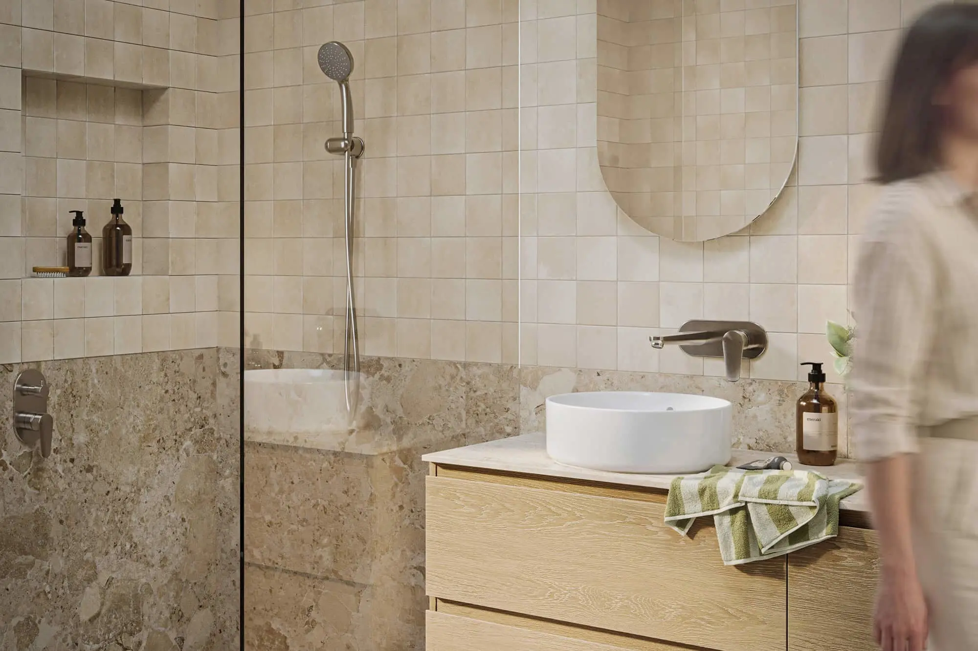 Modern bathroom with beige tiles, glass shower partition, round mirror, and wooden vanity with white vessel sink.