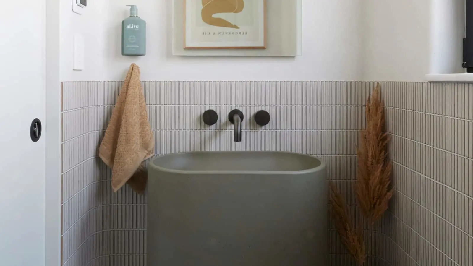 Modern bathroom with green sink, wall-mounted faucet, beige towel, pampas grass, and minimalist artwork above.