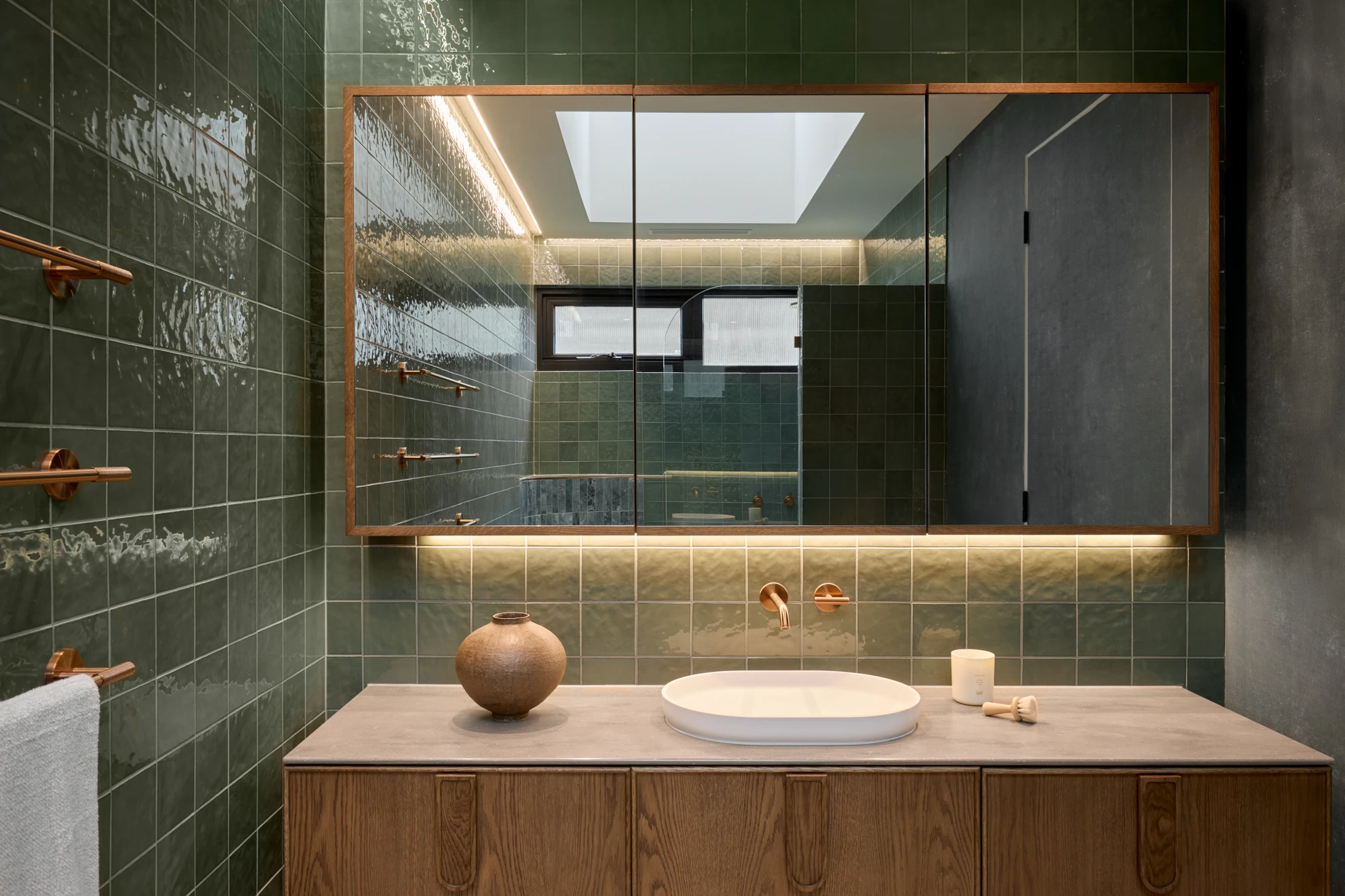 Modern bathroom with green tile walls, wooden vanity, white sink, copper fixtures, and a large mirrored cabinet with lighting.