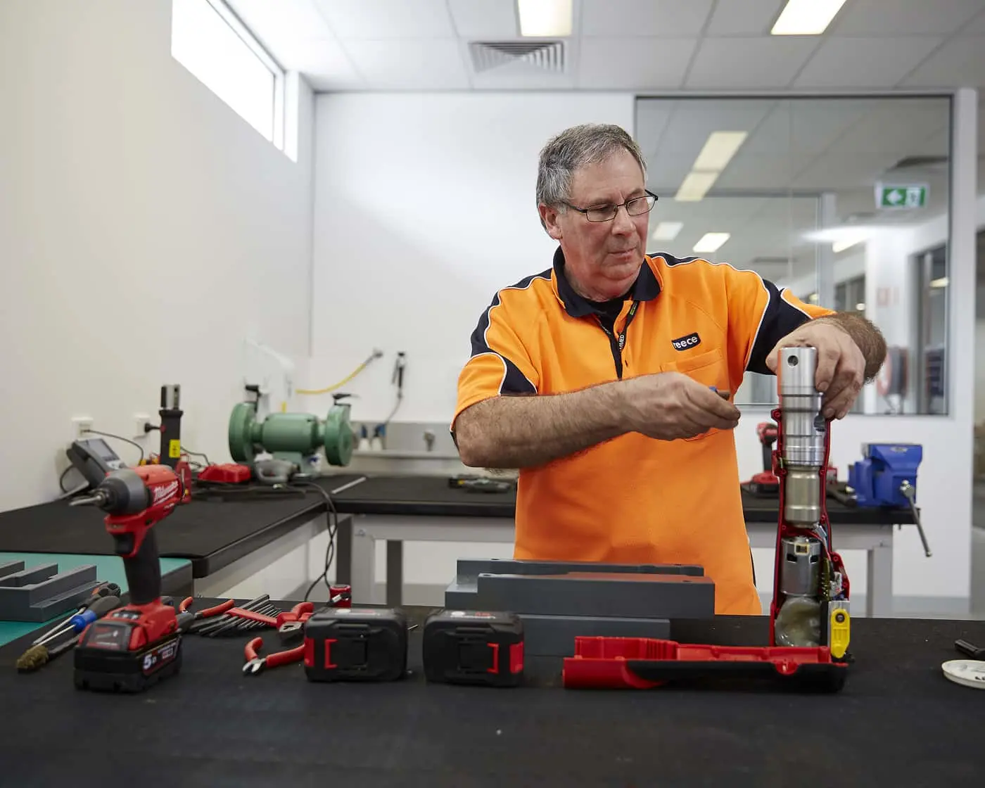 Technician in orange uniform working on mechanical equipment in a workshop with various tools and battery packs on the table.