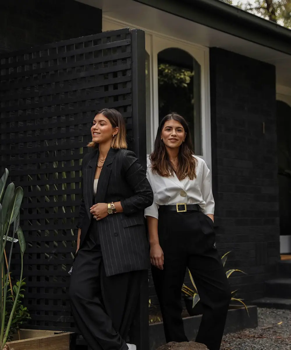 Two women in business attire standing outside a modern home with black lattice screens and white walls.