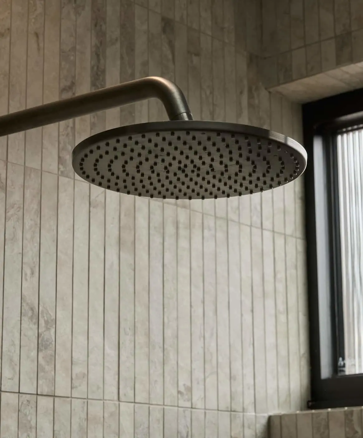 Modern round rainfall showerhead in gunmetal, mounted on a beige tiled bathroom wall next to a window.