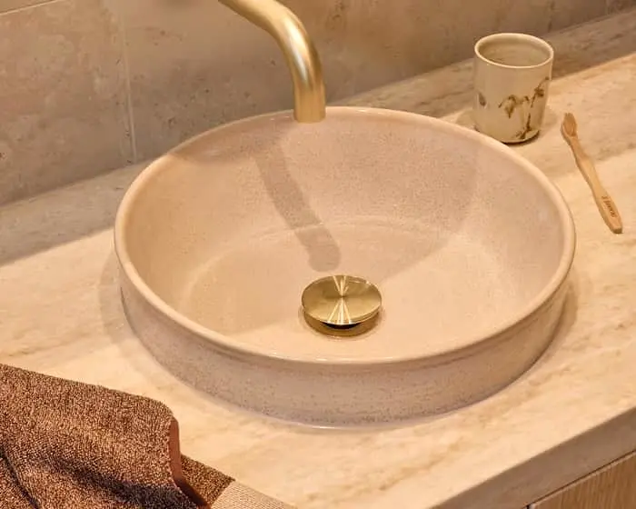 Round beige vessel sink with gold faucet on marble countertop with towel, cup, and toothbrush visible.