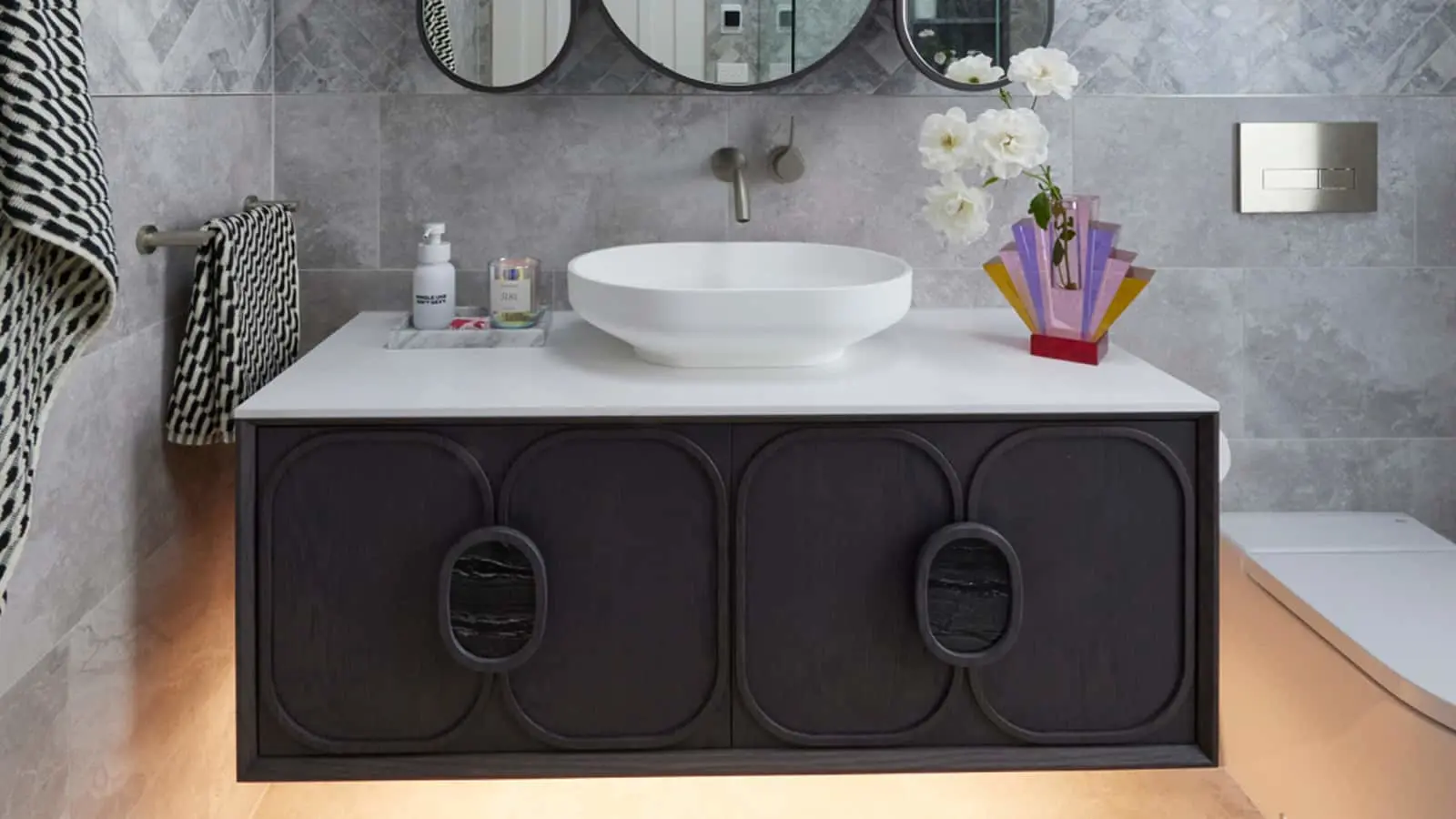 Modern bathroom with white vessel sink on dark cabinet, gray tile walls, round mirrors, and colorful vase with white flowers.