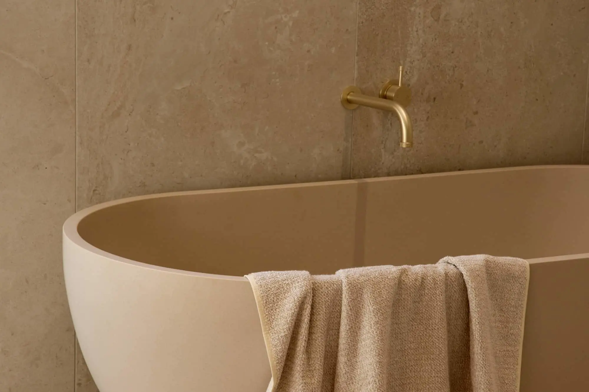 A beige freestanding bathtub with a draped towel, a brass wall-mounted faucet, and beige stone wall tiles in the background.