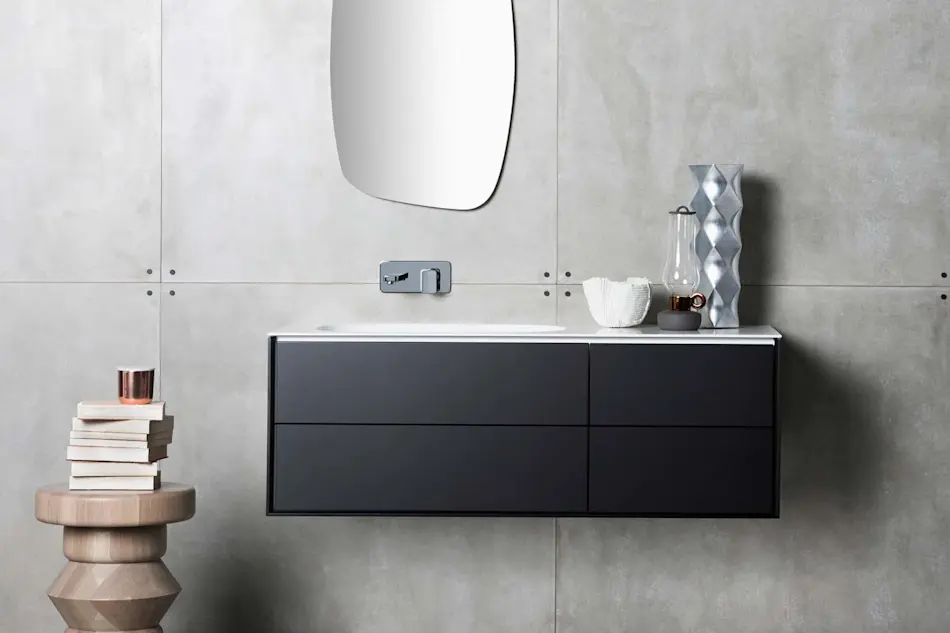 Modern bathroom with floating black vanity, curved mirror, concrete walls, wooden stool with stacked books, and decorative vases.
