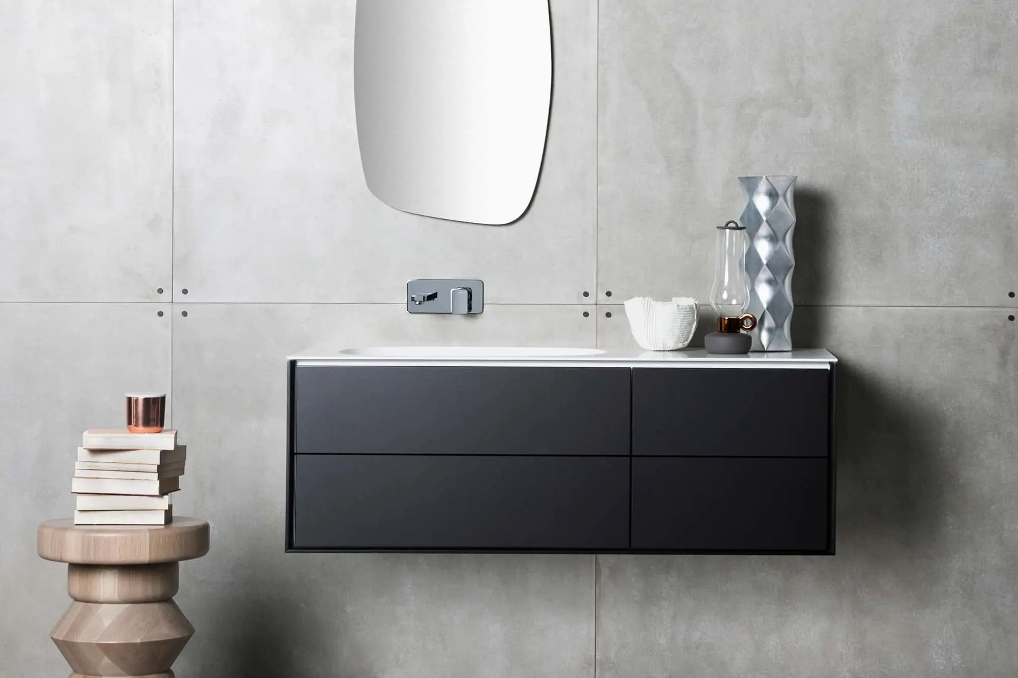 Modern bathroom with floating black vanity, curved mirror, concrete walls, wooden stool with stacked books, and decorative vases.