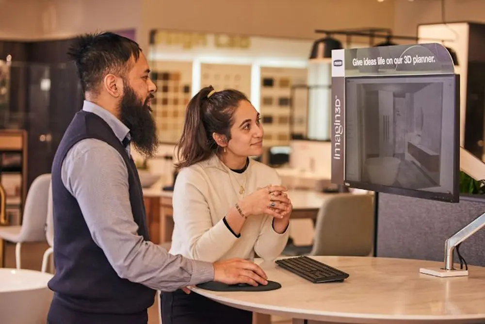Two customers looking at a 3D room planner display in a furniture showroom with "imagine in 3D" text visible.