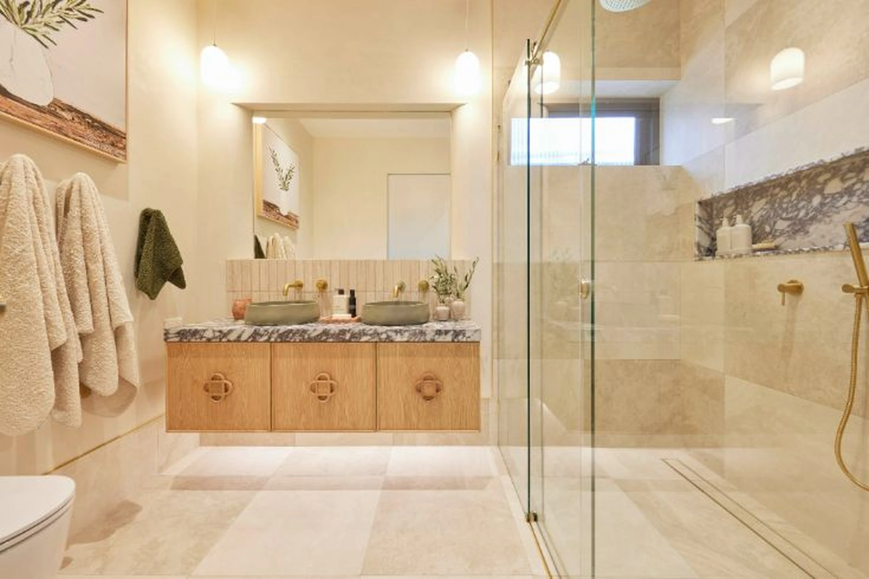 Modern bathroom with wooden vanity, marble countertop, glass shower enclosure, and cream-colored tile in warm lighting.