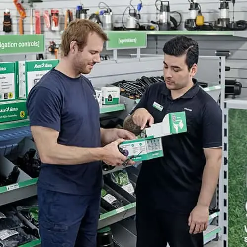 Store employee helping customer select irrigation equipment in hardware store with green product displays.