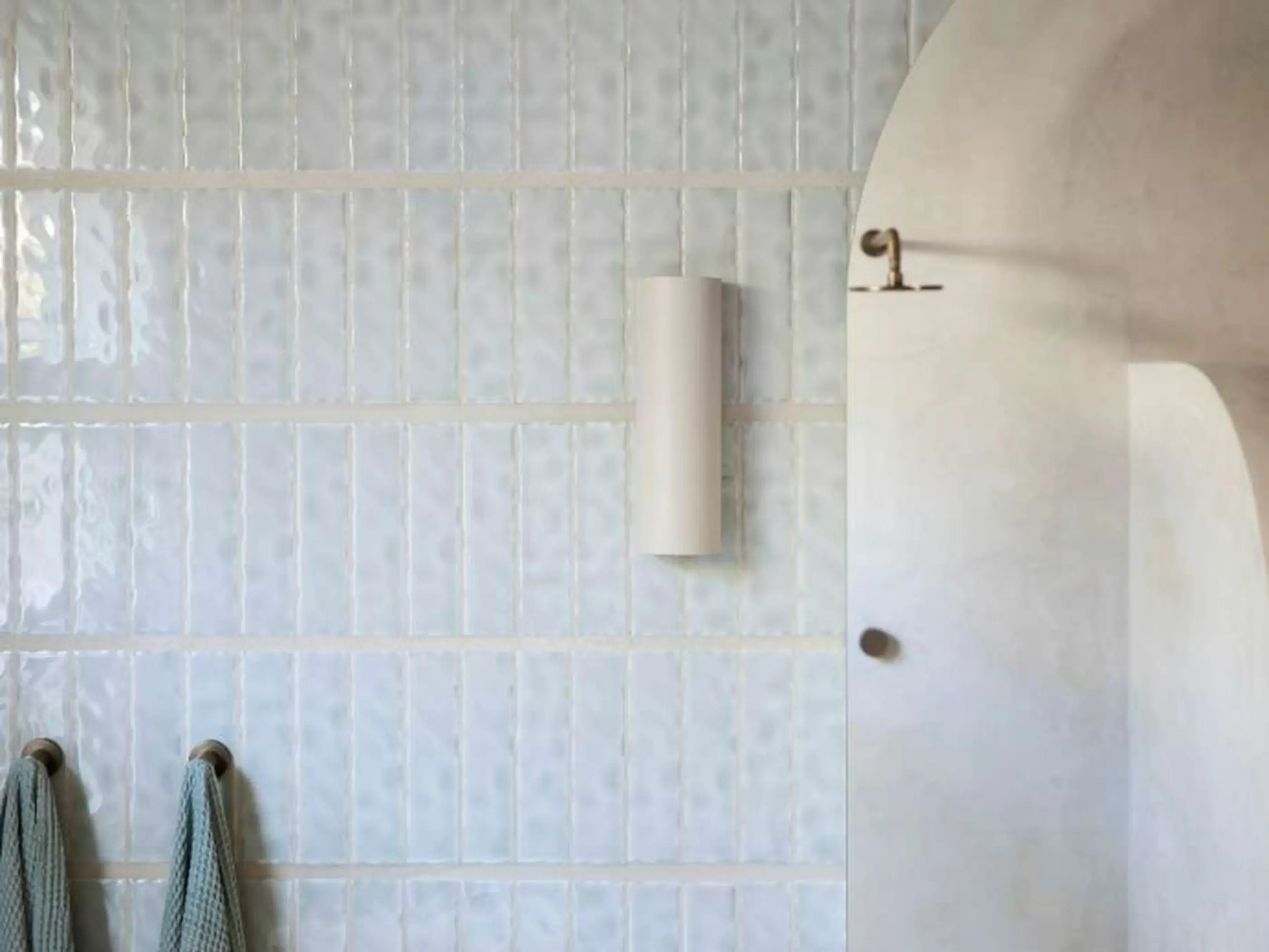 Modern bathroom with light blue vertical tiles, cylindrical wall sconce, shower head, and hanging towels.