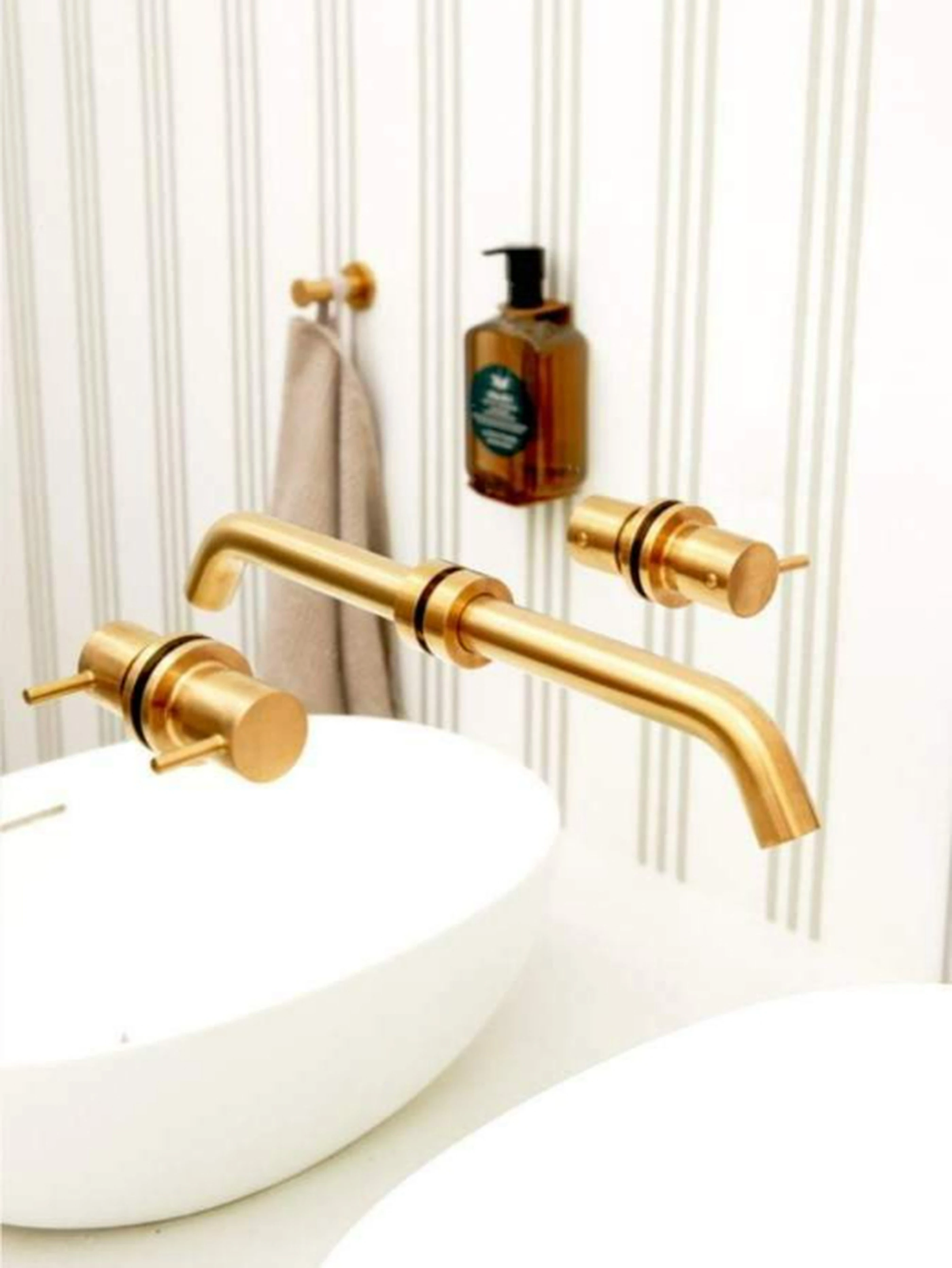 Gold wall-mounted bathroom faucet with white sink, amber soap dispenser, and beige towel on hook against striped wall.